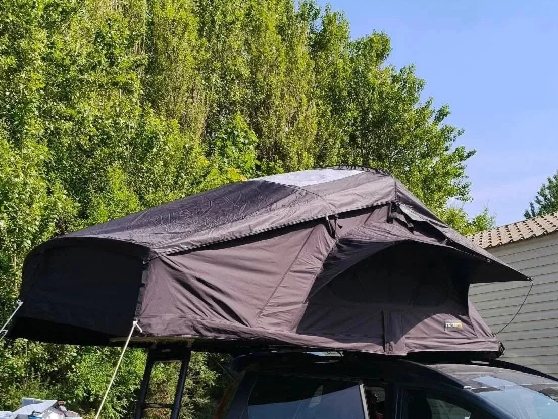 Een zwart daktent bevestigd op het dak van een auto, met een groene bos en een wit gebouw op de achtergrond.