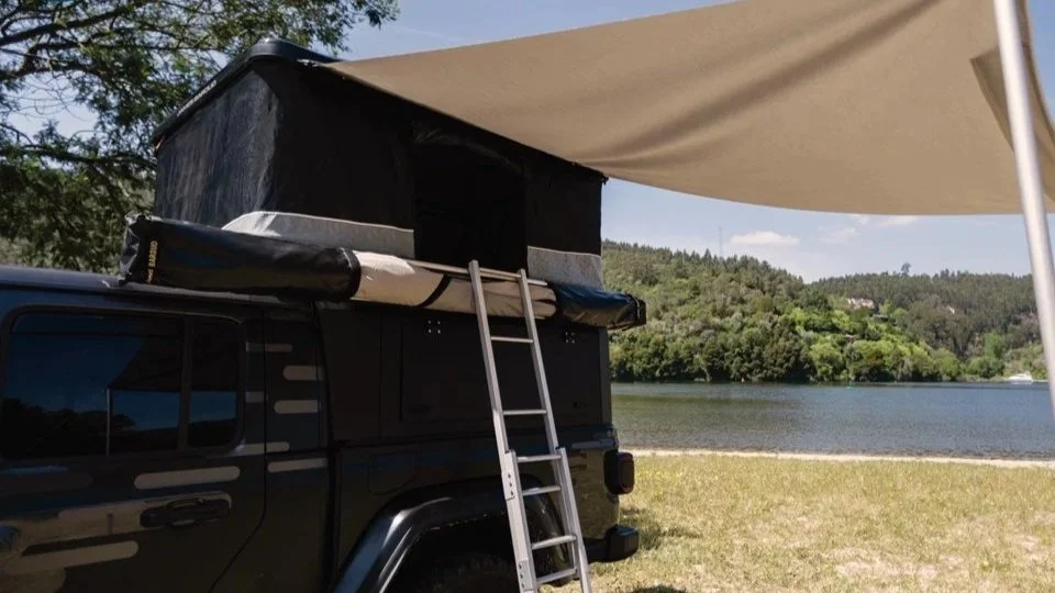 Zwart camper met een daktent op een camping bij een meer, met een ligstoel onder een luifel en een ladder voor de daktent. James Baroud Luifel