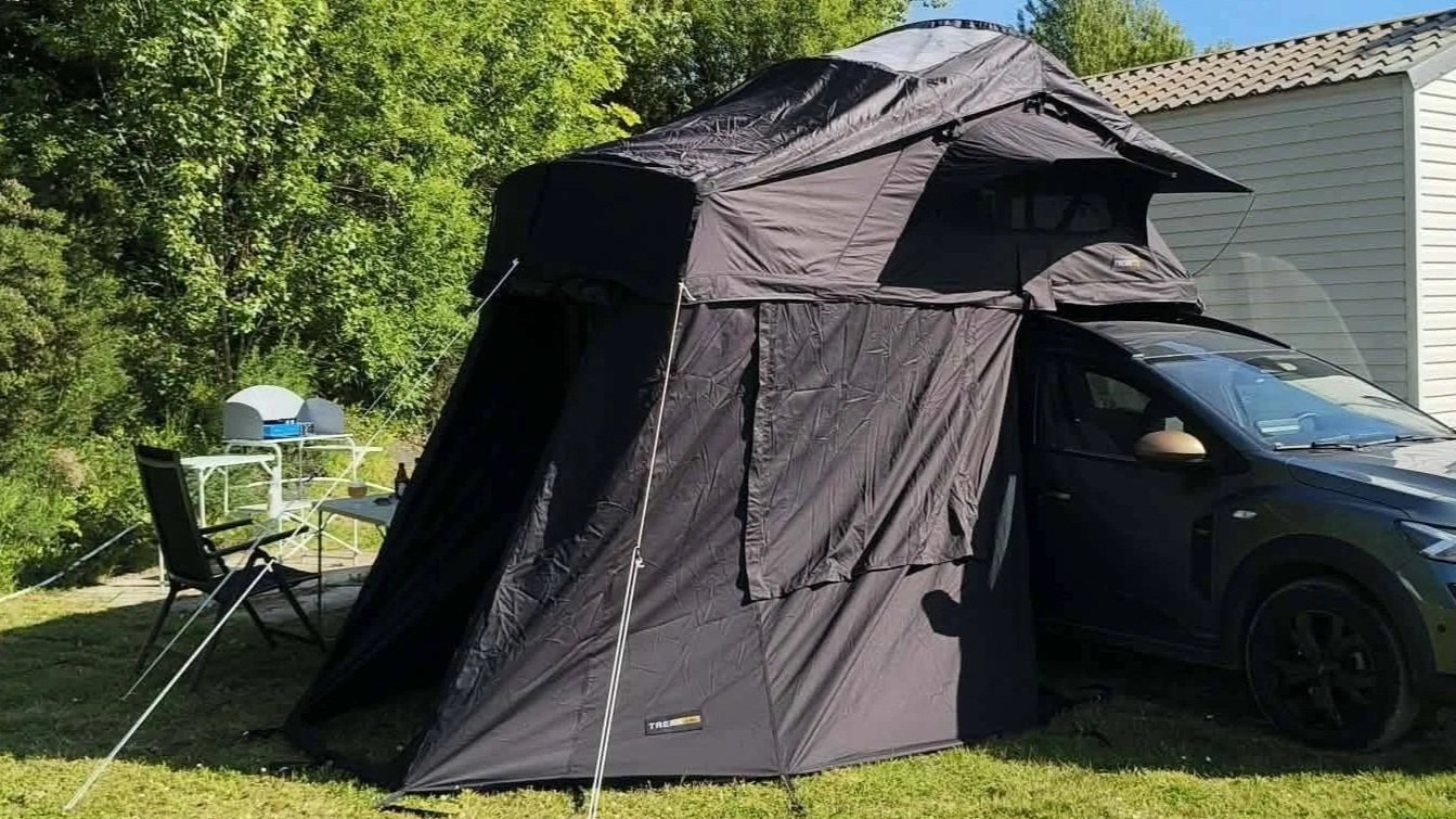 Een auto met een daktent uitgeklapt op een grasveld naast een witte huis. Er staan twee campingstoelen en een kleine tafel met een campingkooktoestel erop.