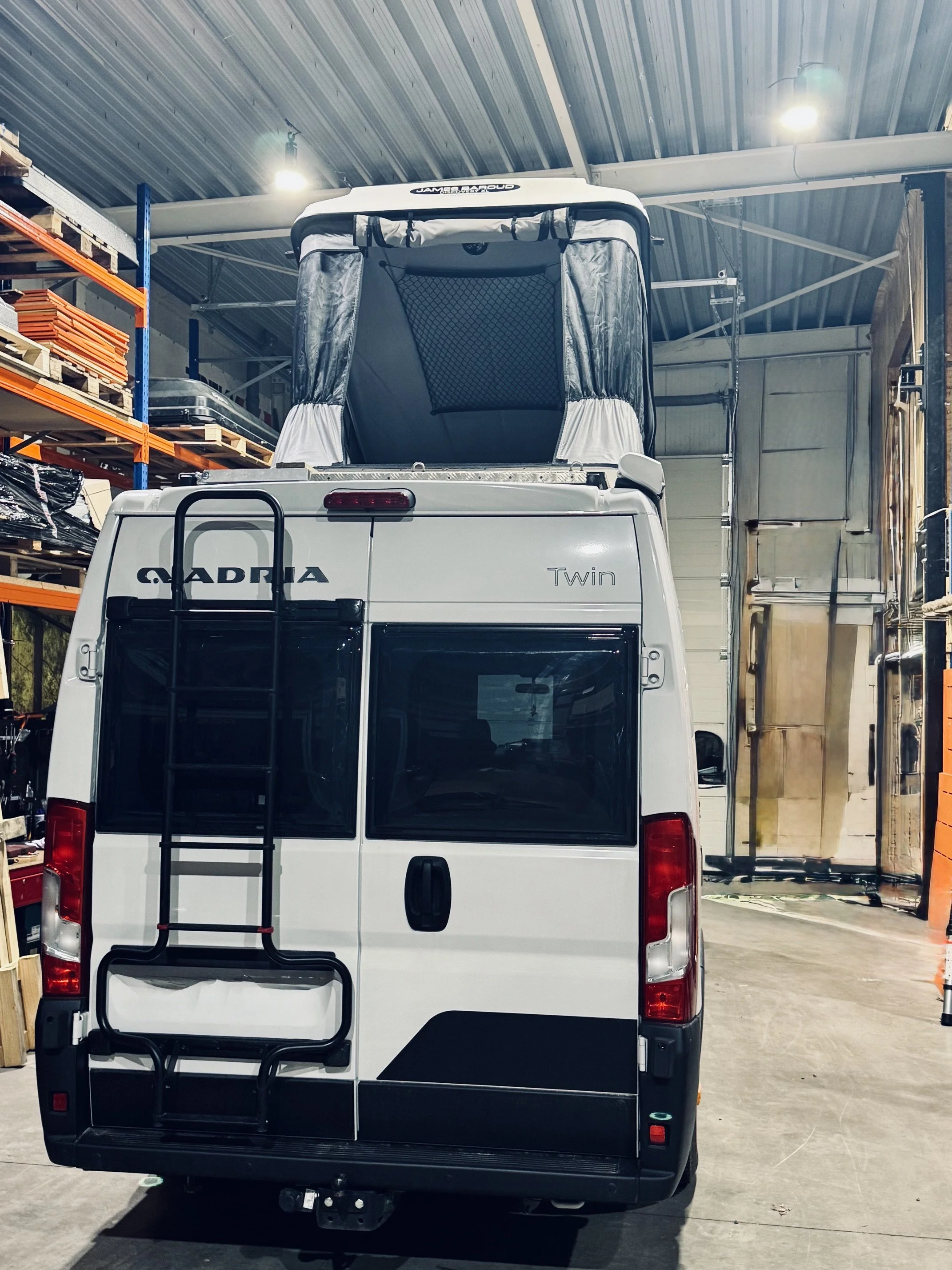 Wit camperbus met fietsendrager, parkeert in een opslagruimte met stalen rekken en houten pallets.