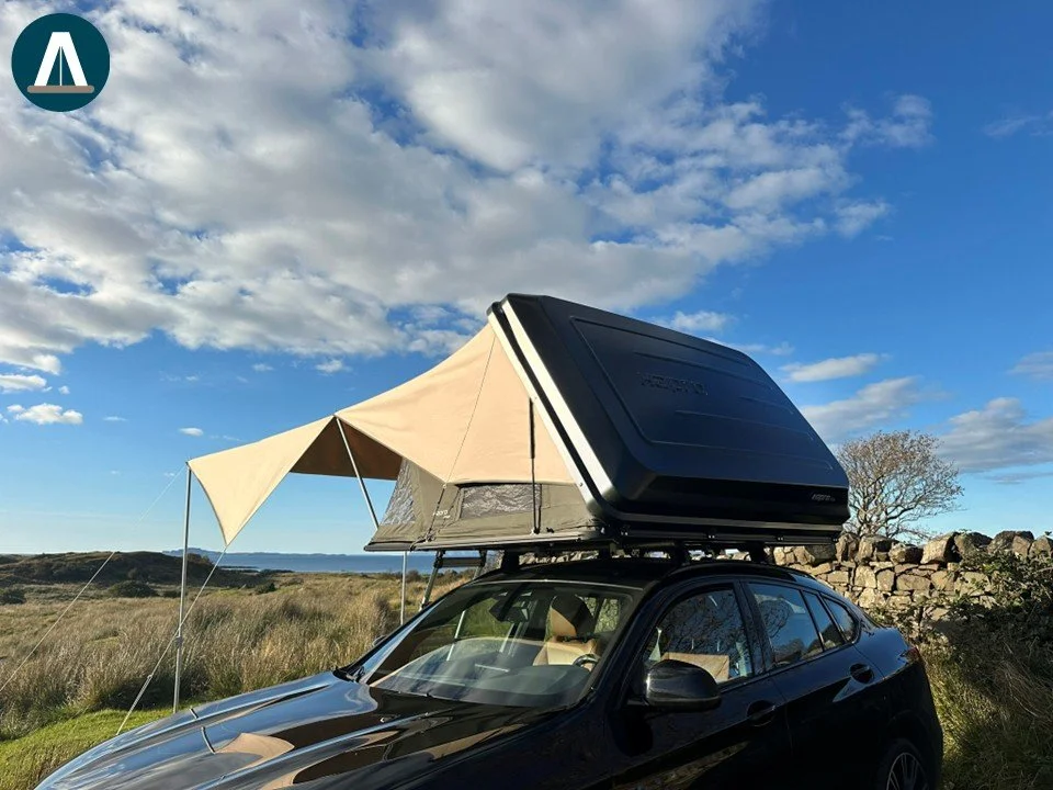 Een zwarte auto met een daktent en een uitklapbare luifel op een open veld onder een met wolken bedekte blauwe hemel.
