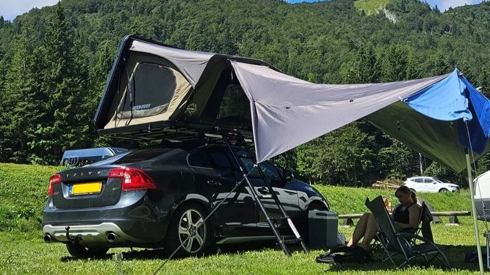 Een zwarte sedan met een daktent en een uitklapbare luifel, een persoon zit in een campingstoel onder de luifel, een groene weide en bomen op de achtergrond. Sheepie Bookara