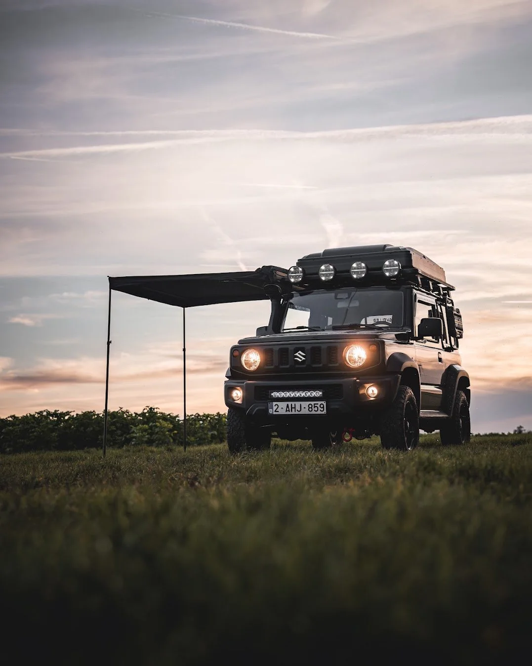 Een zwarte Suzuki Jimny auto met uitrusting, geparkeerd op gras bij zonsondergang, met een zwart afdak en extra lichten op het dak.