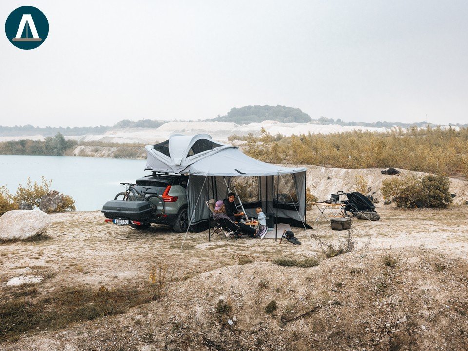 Gezinnen kamperen naast een meer, met een auto en een uitklapbare tent, in een natuurlijke omgeving.
