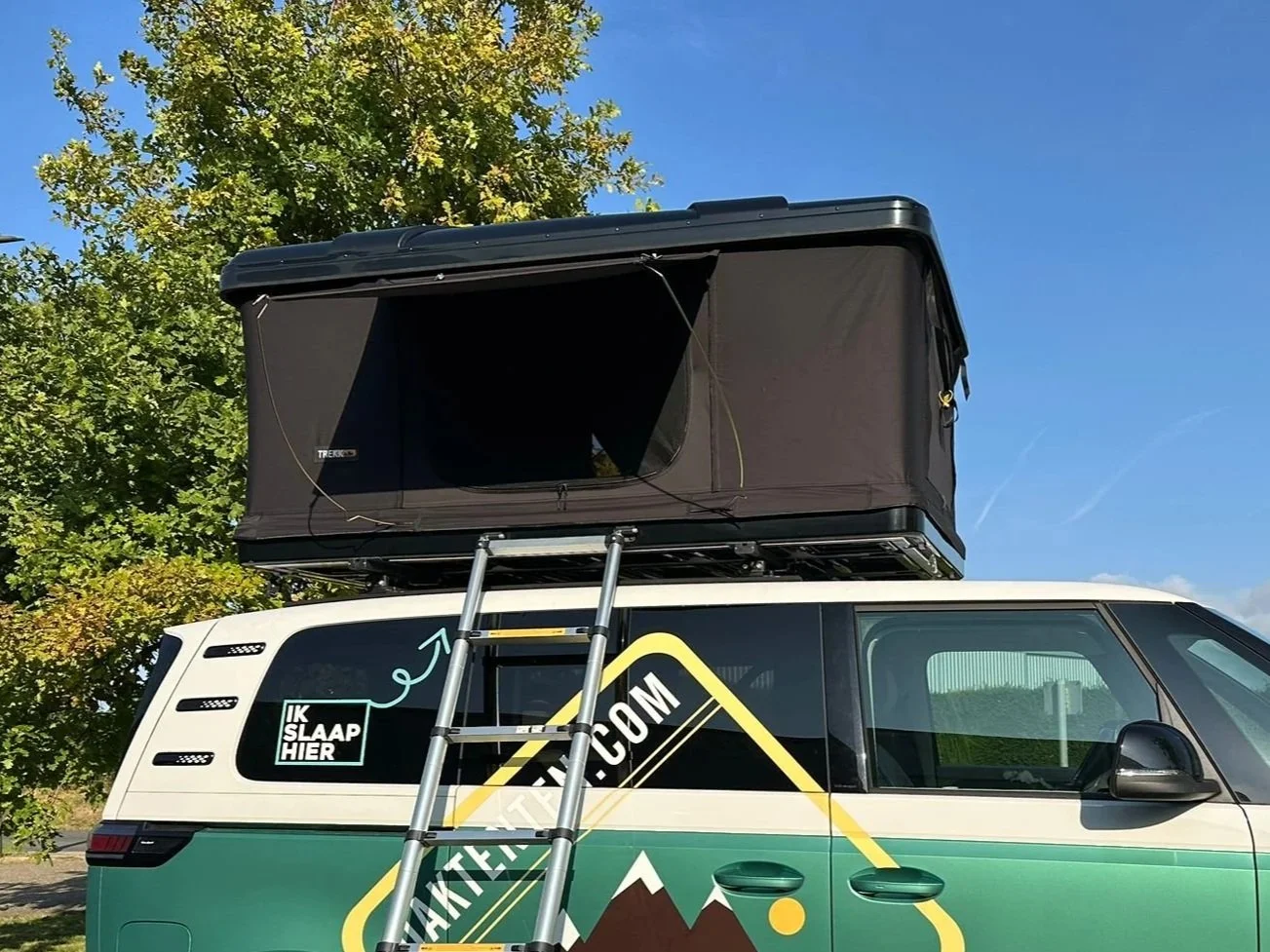 Un camping-car dont la tente de toit est déployée, une échelle appuyée contre le flanc. En arrière-plan, un ciel bleu et des arbres.