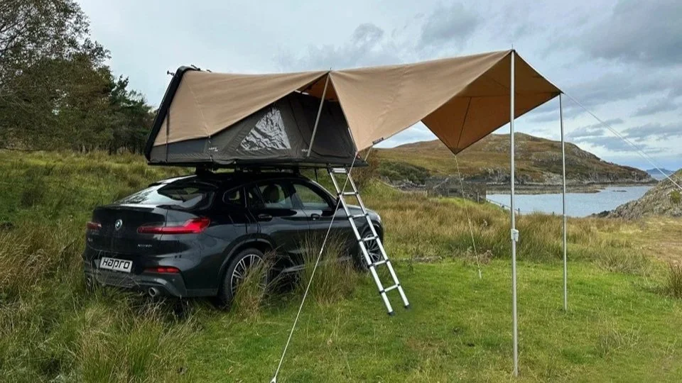 Een zwart SUV met daktent en luifel in een groene, landelijke omgeving met water en heuvels op de achtergrond. Hapro Vista