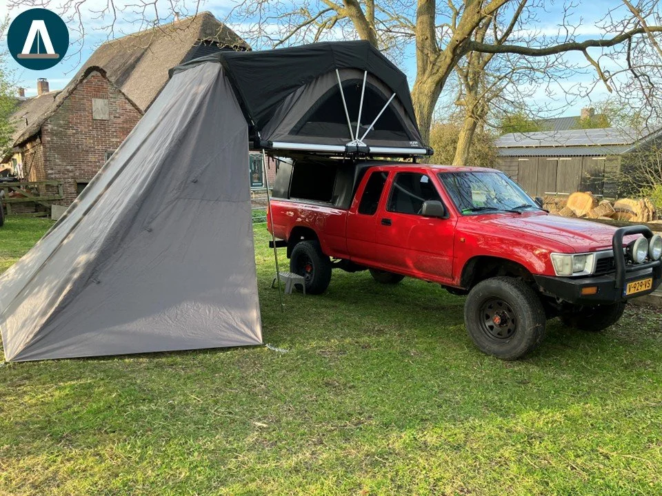 Een rode pickup truck met een kampeeruitrusting, waaronder een daktent en een voortent, in een tuin met gras en bomen.