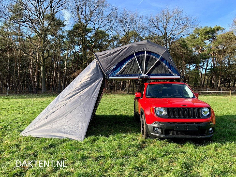 Een rode Jeep met een tentopbouw die uit de auto is uitgeklapt, gelegen op een grasveld met bomen op de achtergrond.