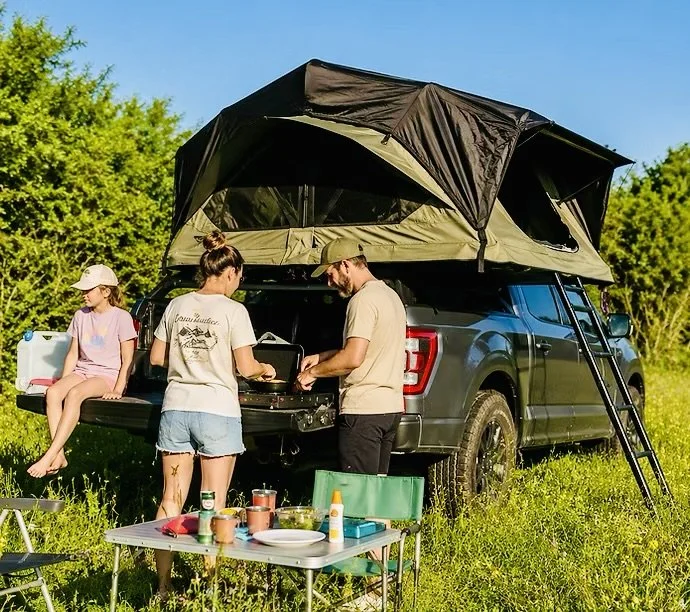 Een gezin telt drie personen, een vrouw, een man en een meisje, die genieten van een kampeeruitje naast een geparkeerde camper met een daktent, met een tafel vol eten en drinken op een grasveld onder een heldere blauwe lucht.