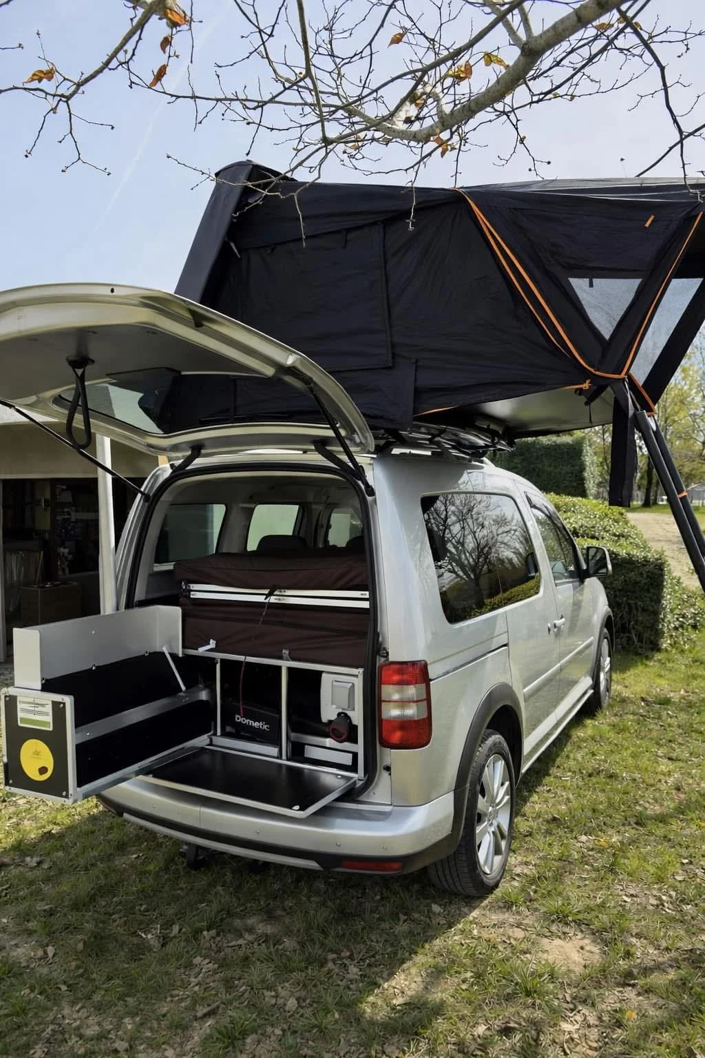 Een zilverkleurige auto met een uitklapbare tapijt op het dak en een uitklapbare keuken aan de achterzijde, geparkeerd op gras. Bomen en een huis zijn zichtbaar in de achtergrond.