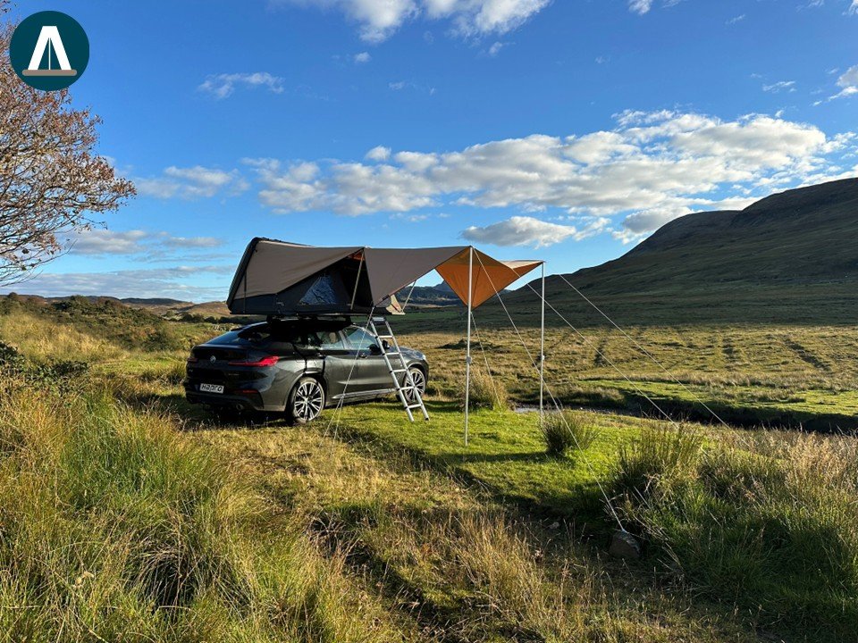 Een auto met een daktent en een voortent op een grasveld in een bergachtig landschap onder een blauwe hemel met wolken.