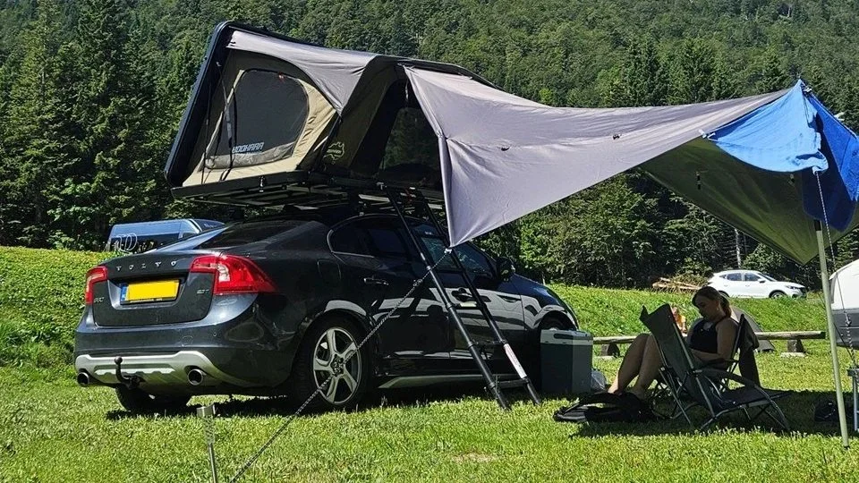Een zwarte Volvo stationwagen met een ingebouwd tentdak op een grasveld in een bosrijke omgeving, met een vrouw die in een ligstoel zit onder een grote luifel.