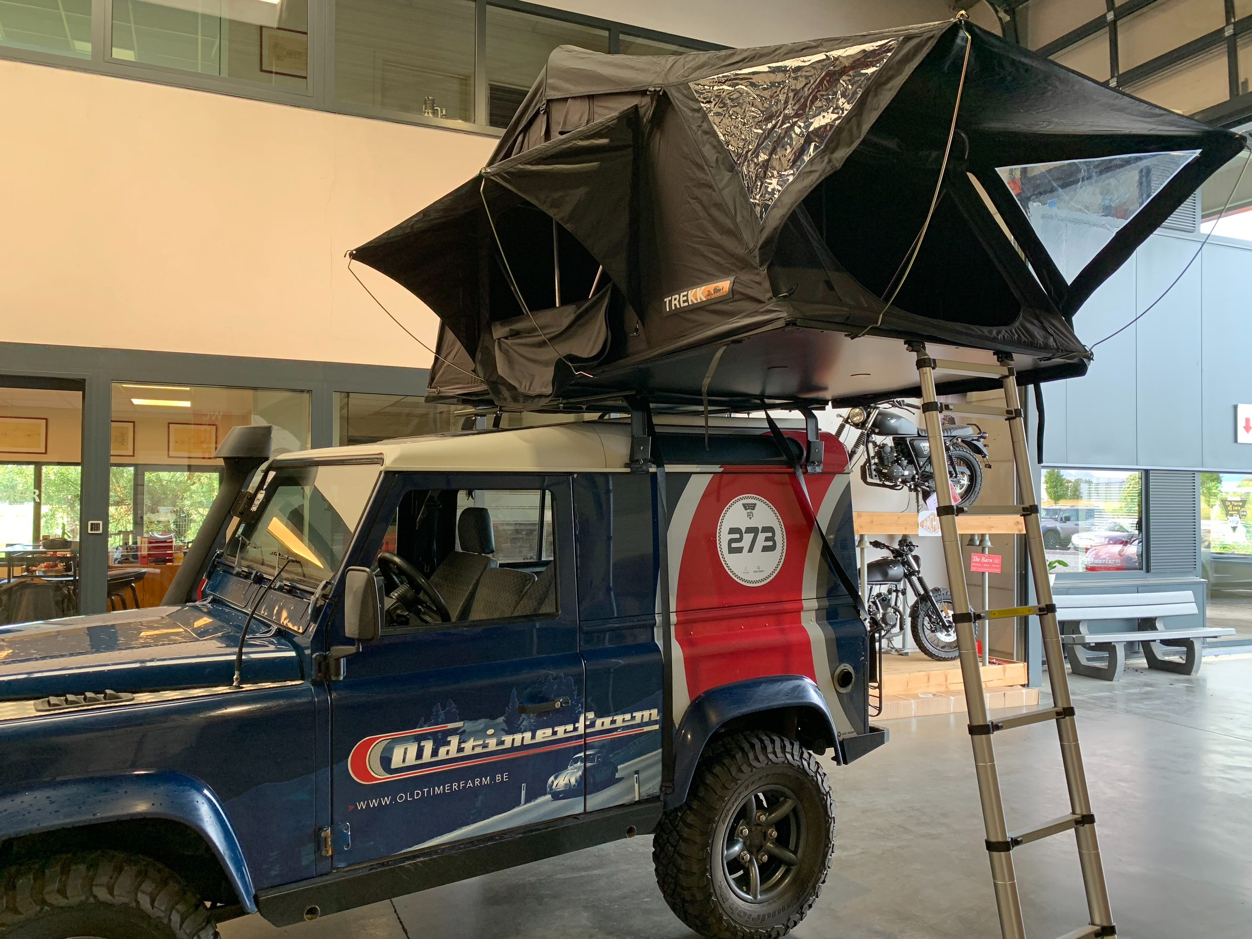Een oude blauwe Land Rover met een dakdrone en twee fietsen op het dak, binnen een gebouw met grote ramen.