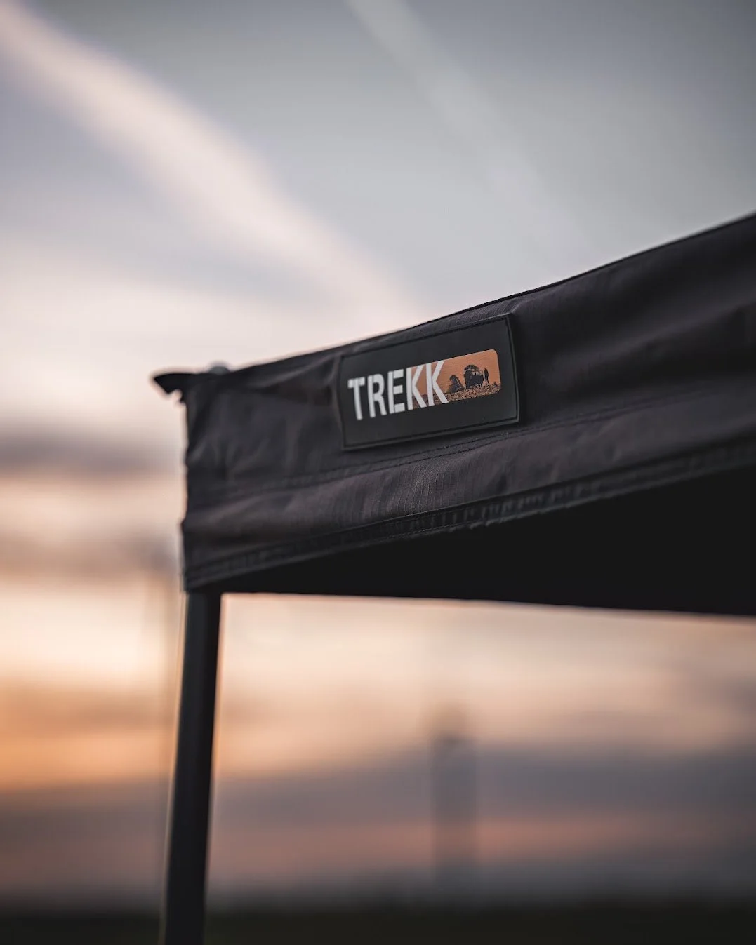 Een close-up van een zwart krijtbord met het logo en de naam 'TREKK' en een silhouet van een landschap met kamelen, tegen een sunsetachtergrond met een gevlekte hemel.