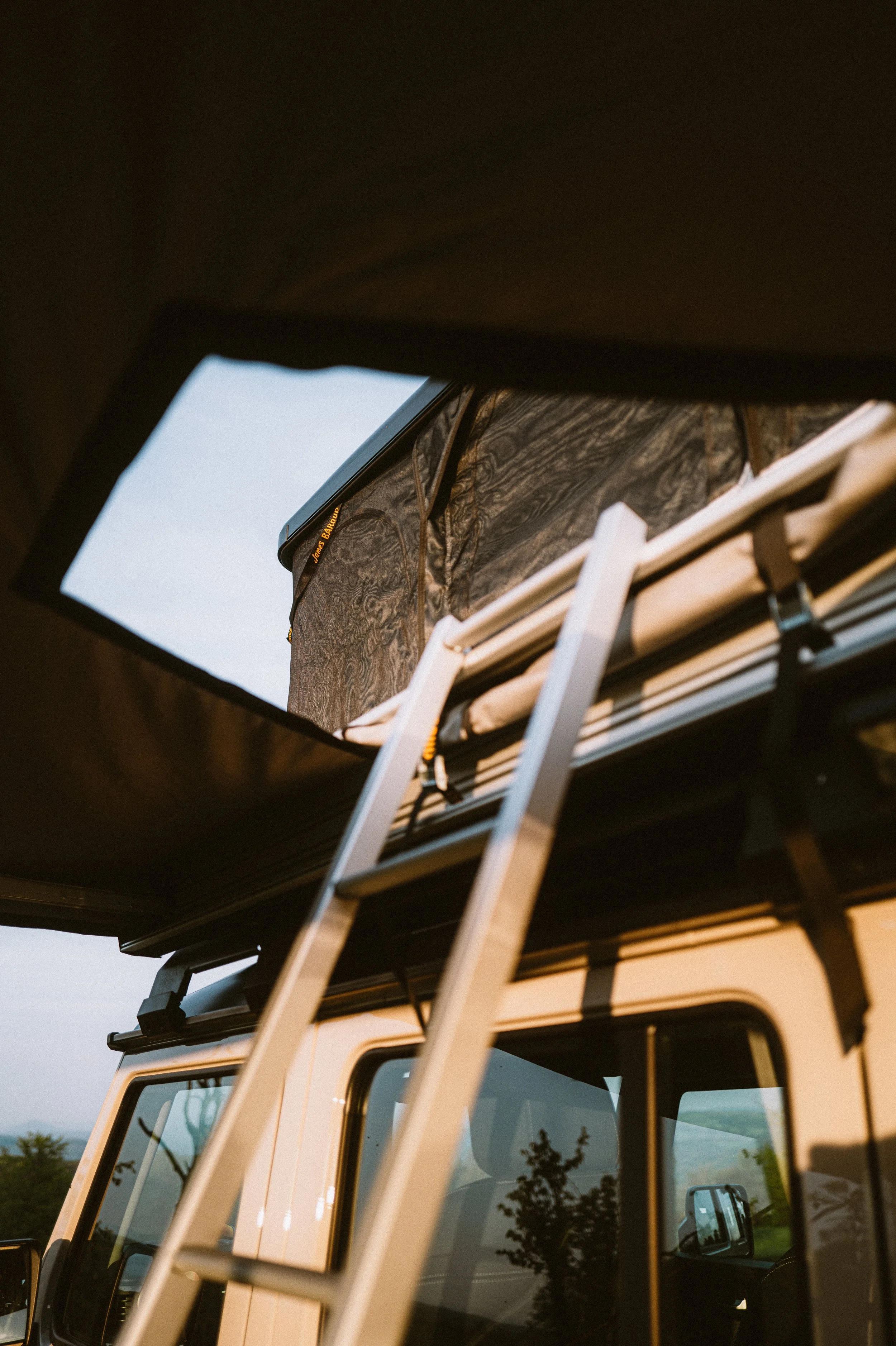 Een foto genomen vanuit onder de dakrand van een camper, gericht op dakkoffer, ladder en de bovenkant van de camper met ramen en spiegel. Het is vroeg in de ochtend of late in de avond, met een rustige omgeving en de reflectie van een boom in de ramen.