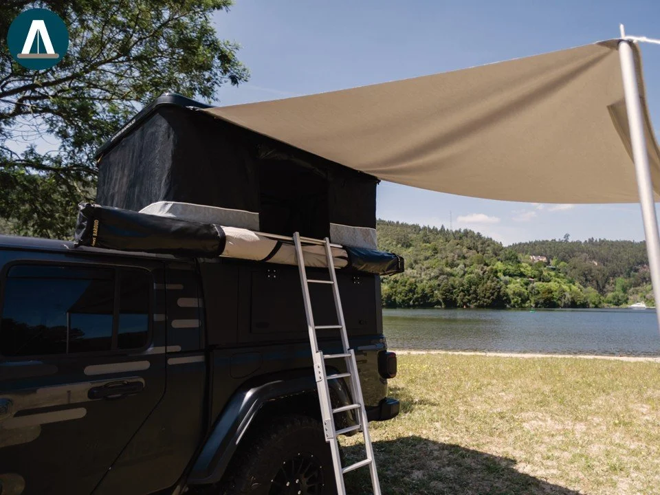 Een zwarte camper met een opbouwtent en een ladder aan de zijkant, naast een meer met groene heuvels op de achtergrond en een blauwe lucht. James Baroud Luifel