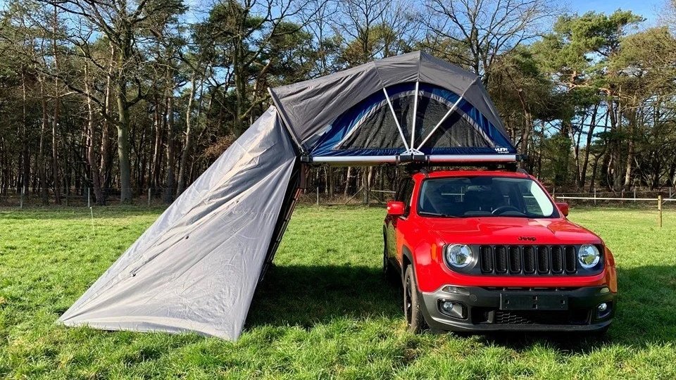 Een rode Jeep Cherokee met een tent bevestigd bovenop, die uitstrekt over een grasveld met bomen op de achtergrond. Sheepie Yuna Jimba