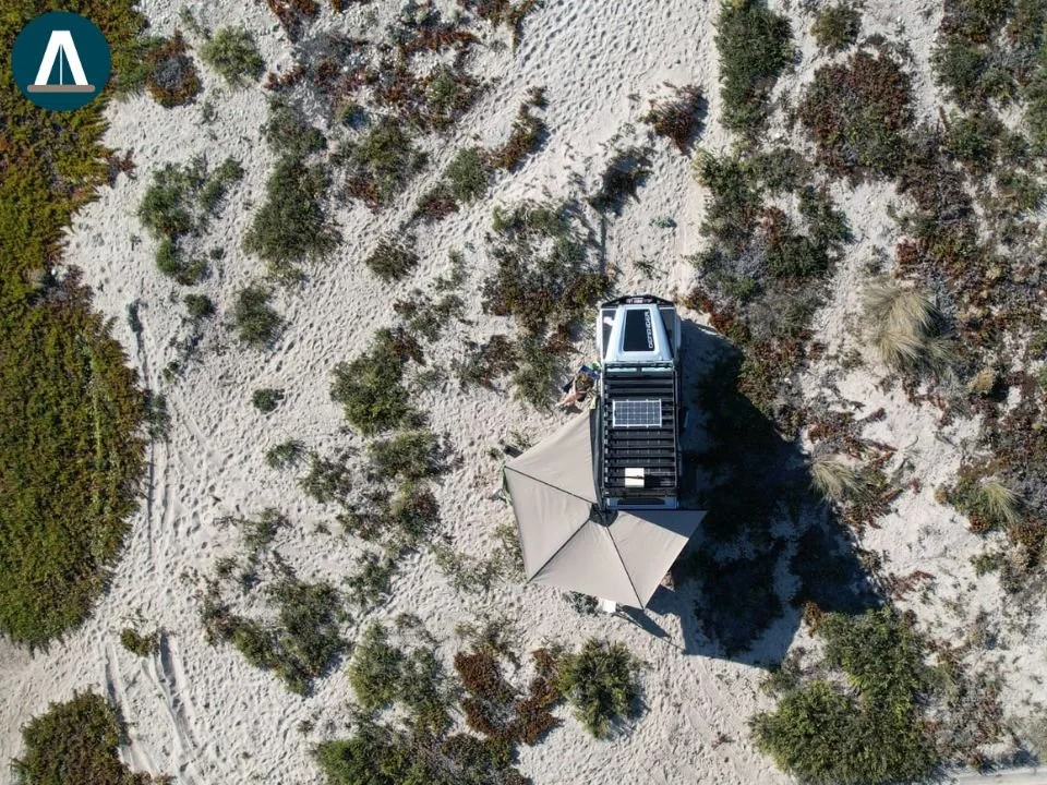 Een dronefoto van een camper met een zonnepaneel en een parasol op een zandstrand met verspreide struiken en planten.