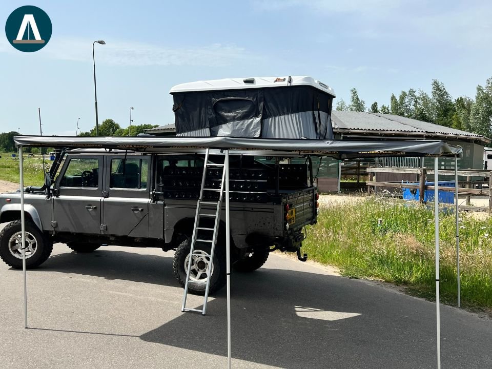 Een donkergrijze terreinwagen met een daktent en een kano of kajak op het dak. De auto staat geparkeerd op een weg naast een veld met gras en een schuur in de achtergrond. James Baroud Luifel