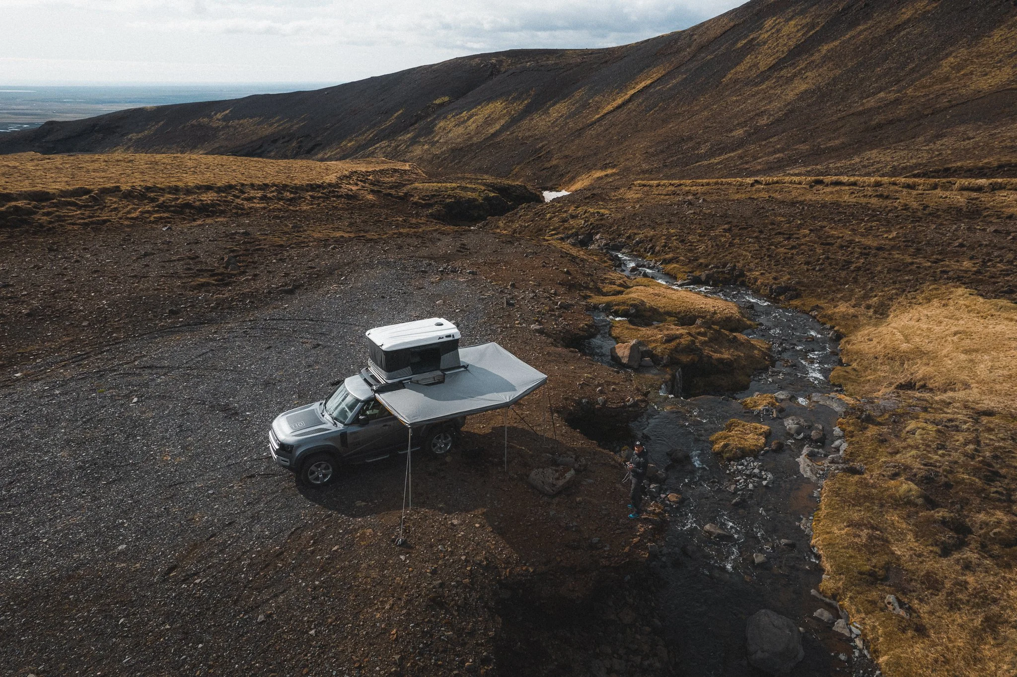 Een camper staat op een rotsachtig terrein naast een kabbelend beekje, met uitgestrekte bergen en een bewolkte hemel op de achtergrond in een bergachtig landschap. James Baroud Luifel