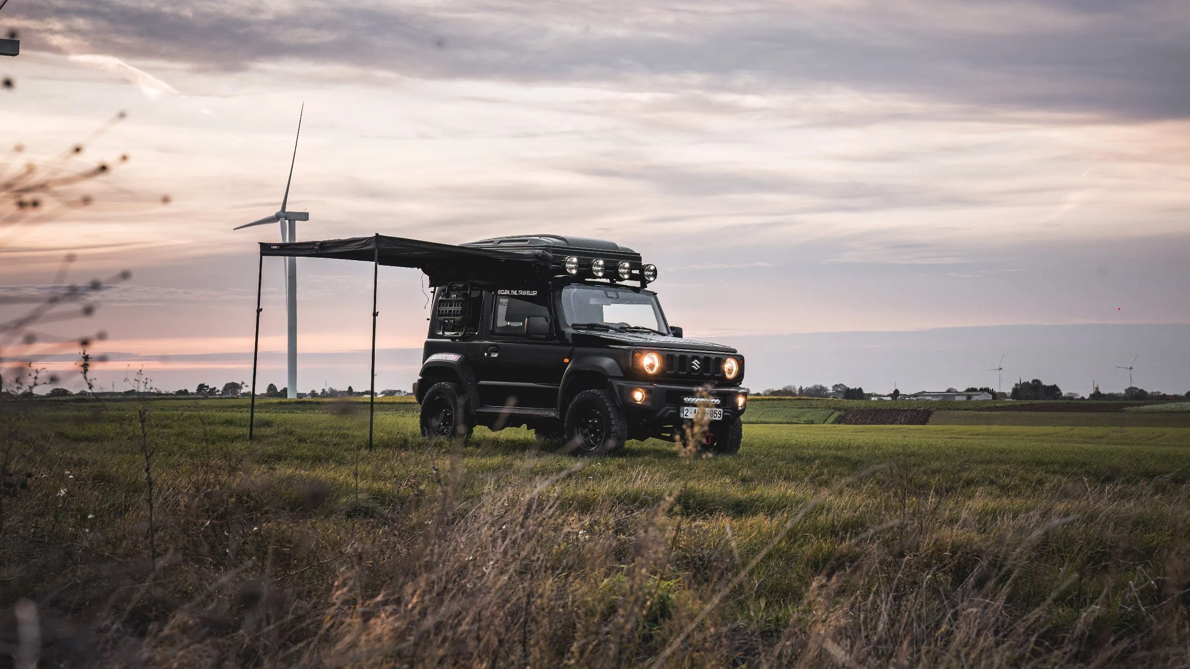 Een zwart off-road voertuig met een afdak en vier koplampen staat op een grasveld tijdens zonsondergang, met windturbines op de achtergrond. Luifel LED