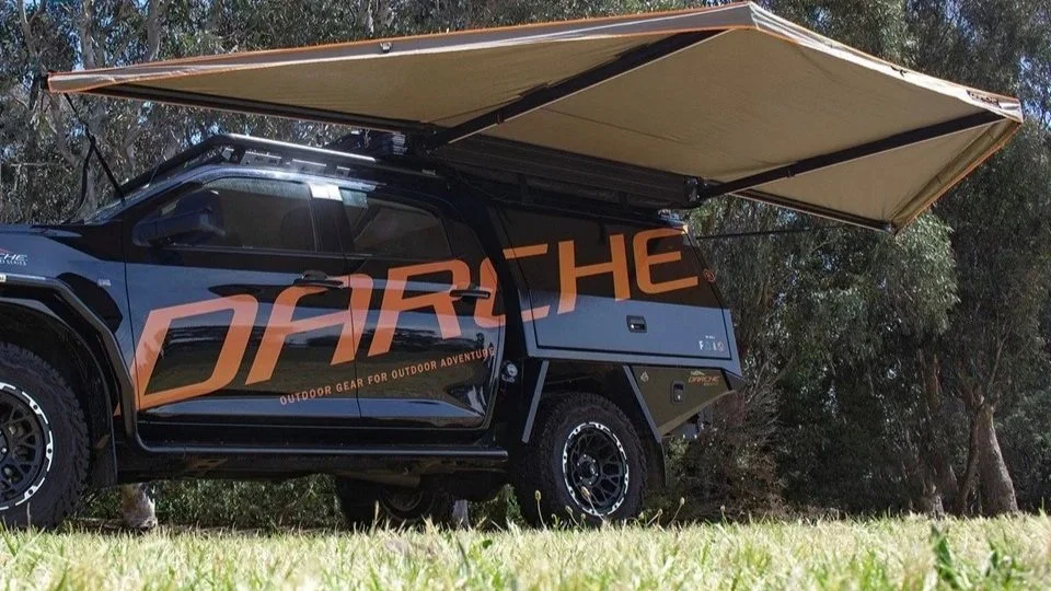 Een terreinwagen met grote banden en een opvouwbare dakborder, met de tekst 'DARCHE' en 'outdoor gear for outdoor adventures' erop, geparkeerd op een grasveld met bomen op de achtergrond.