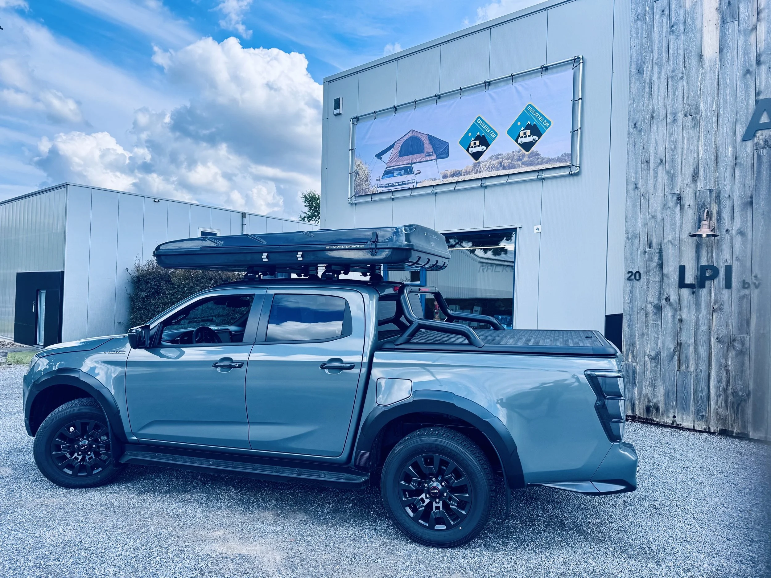 Een blauwe pick-up truck met een dakkoffer op het dak staat voor een modern gebouw met een reclamebord dat een tent en auto afbeeldt, onder een blauwe hemel met wolken.