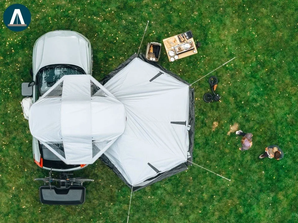 Luchtfoto van een kamp met een witte tent, een pick-up truck en mensen die praten op een grasveld.