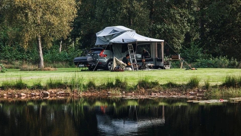 een kampeerplek naast een water met een auto en een camouflage tent, met mensen die binnen en buiten de tent zitten, omringd door bomen.