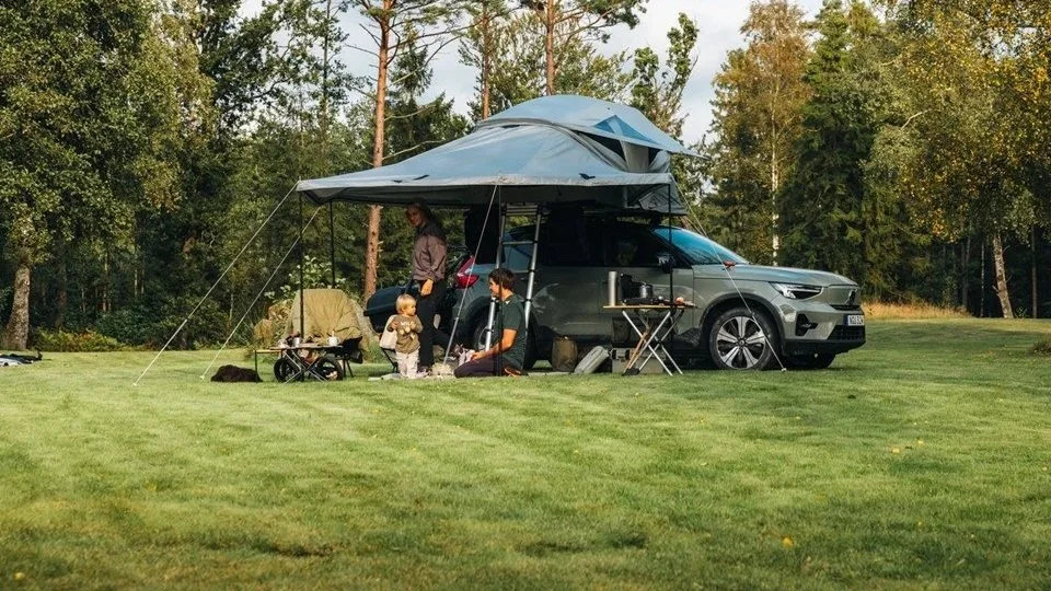 Gezinnen die kamperen op een open grasveld met een auto en een tent. Er zijn twee volwassenen en twee kinderen, en er staat een tafel met kookbenodigdheden. Thule Approach 2 luifel
