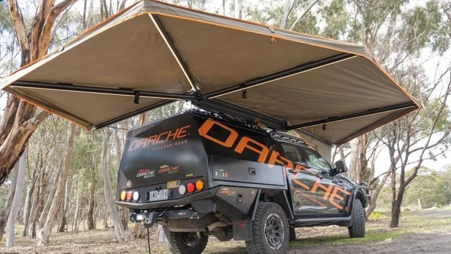 Un camping-car noir portant l'inscription « DARCHE » en grosses lettres orange sur le côté, sous un grand auvent rabattable, garé dans une zone boisée.