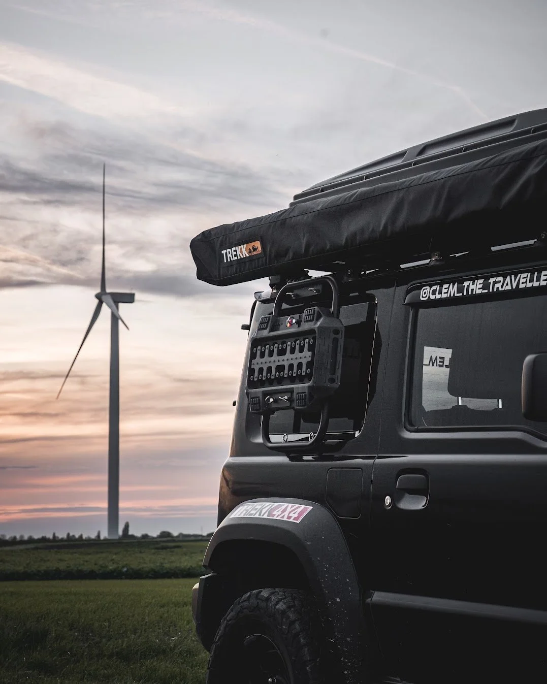 Zwart off-road voertuig met uitrusting voor camping en buitenactiviteiten, op een veld met windturbine op de achtergrond, tijdens zonsondergang. Luifel LED