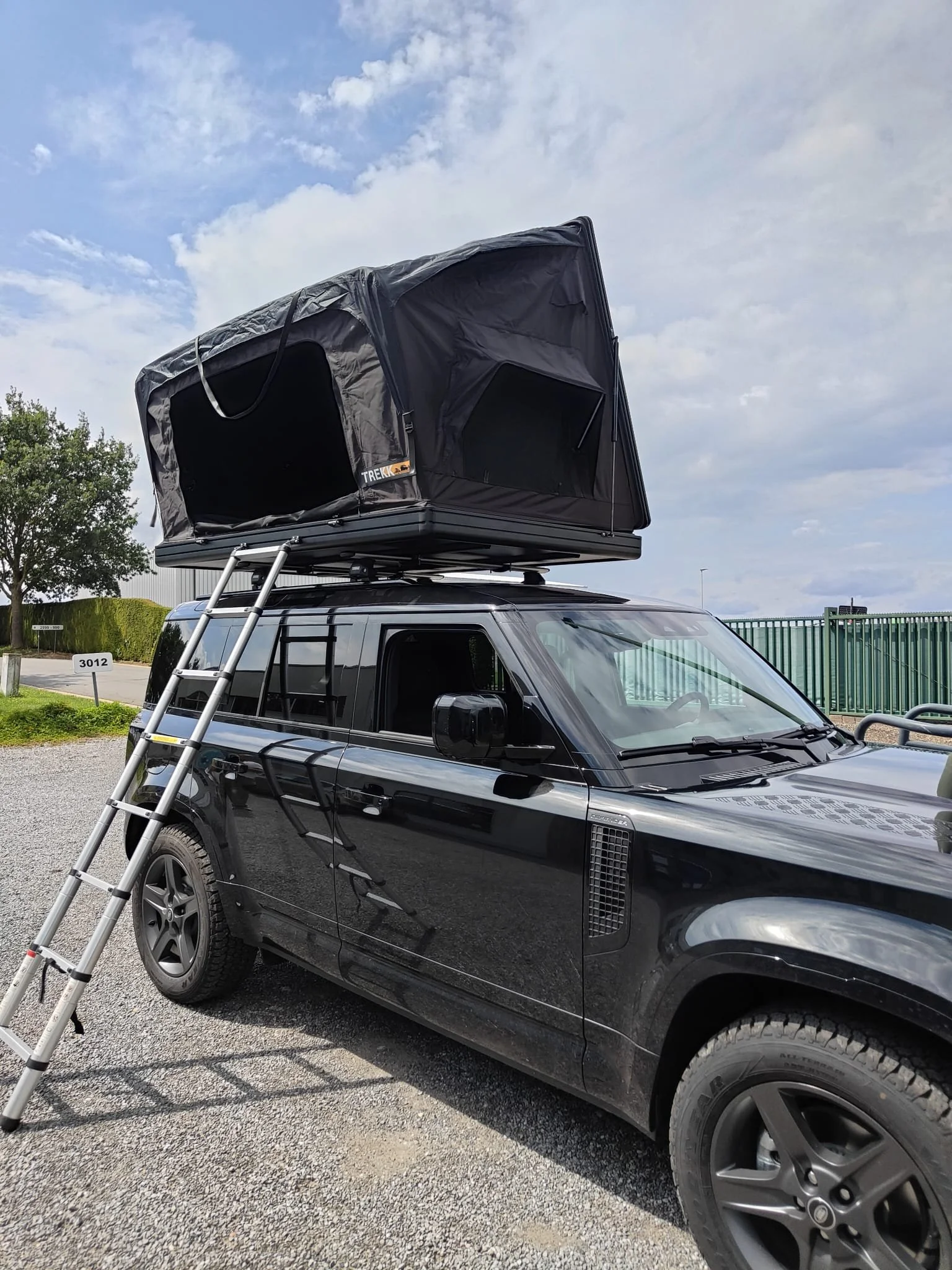 Zwarte off-road voertuig met daktent en ladder, geparkeerd op grind, onder een bewolkte hemel.