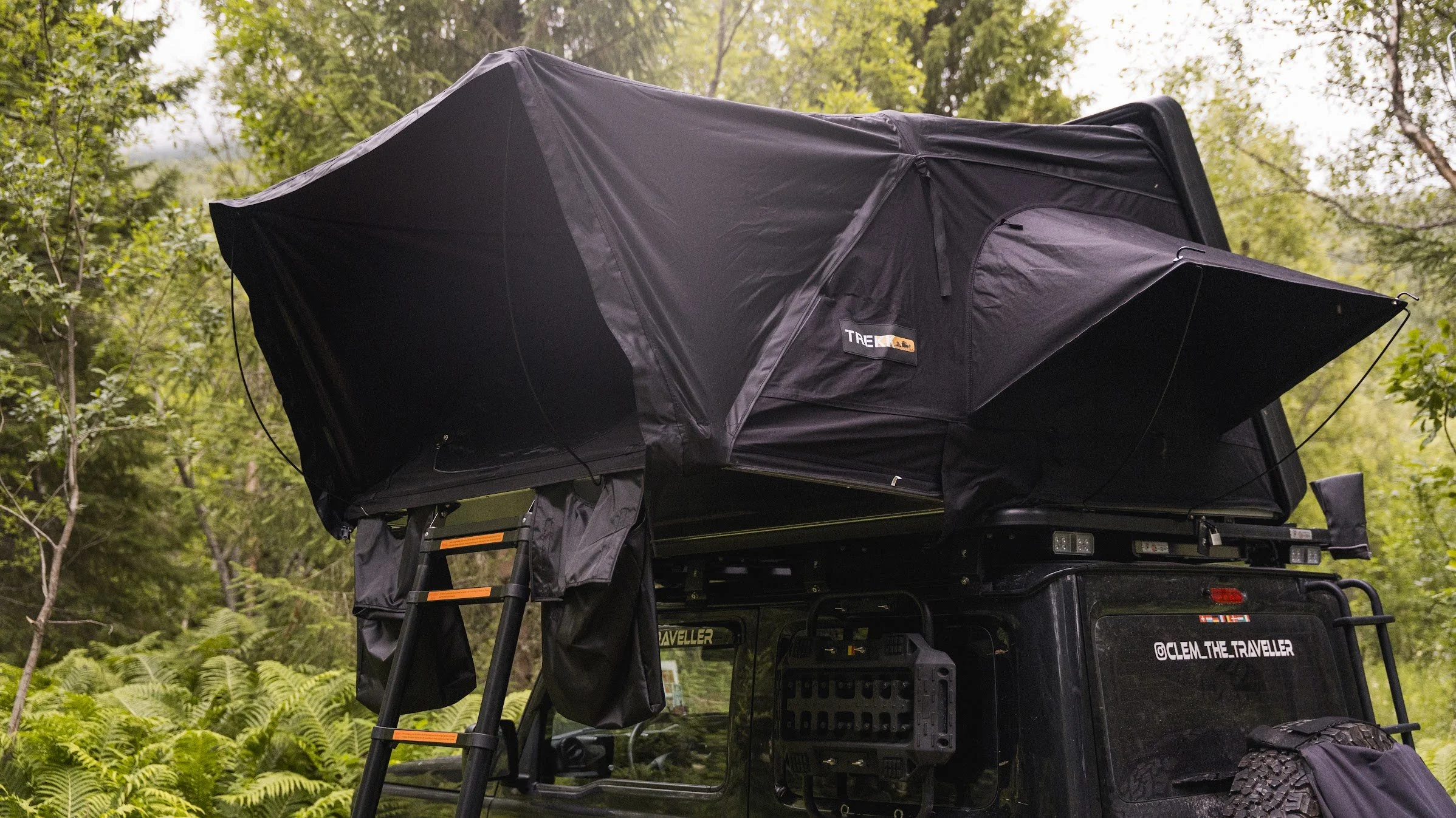 Een zwart kampeerbusje met een opklapbaar tentdak, geplaatst in een bosrijk gebied met groene bomen en planten.