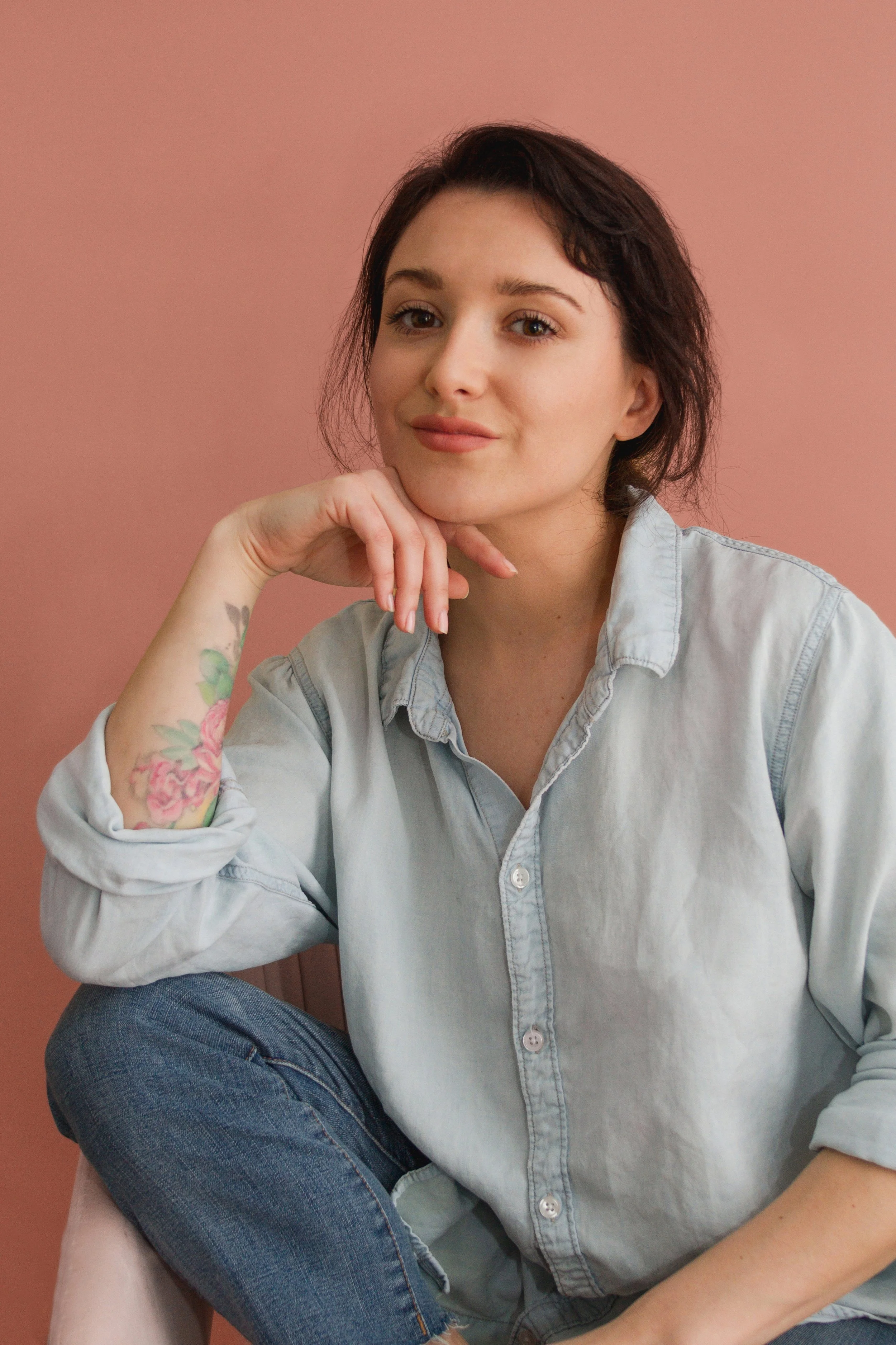 A woman with short dark hair, wearing a light denim shirt and blue jeans, sitting against a pink background with her chin resting on her hand, looking at the camera.