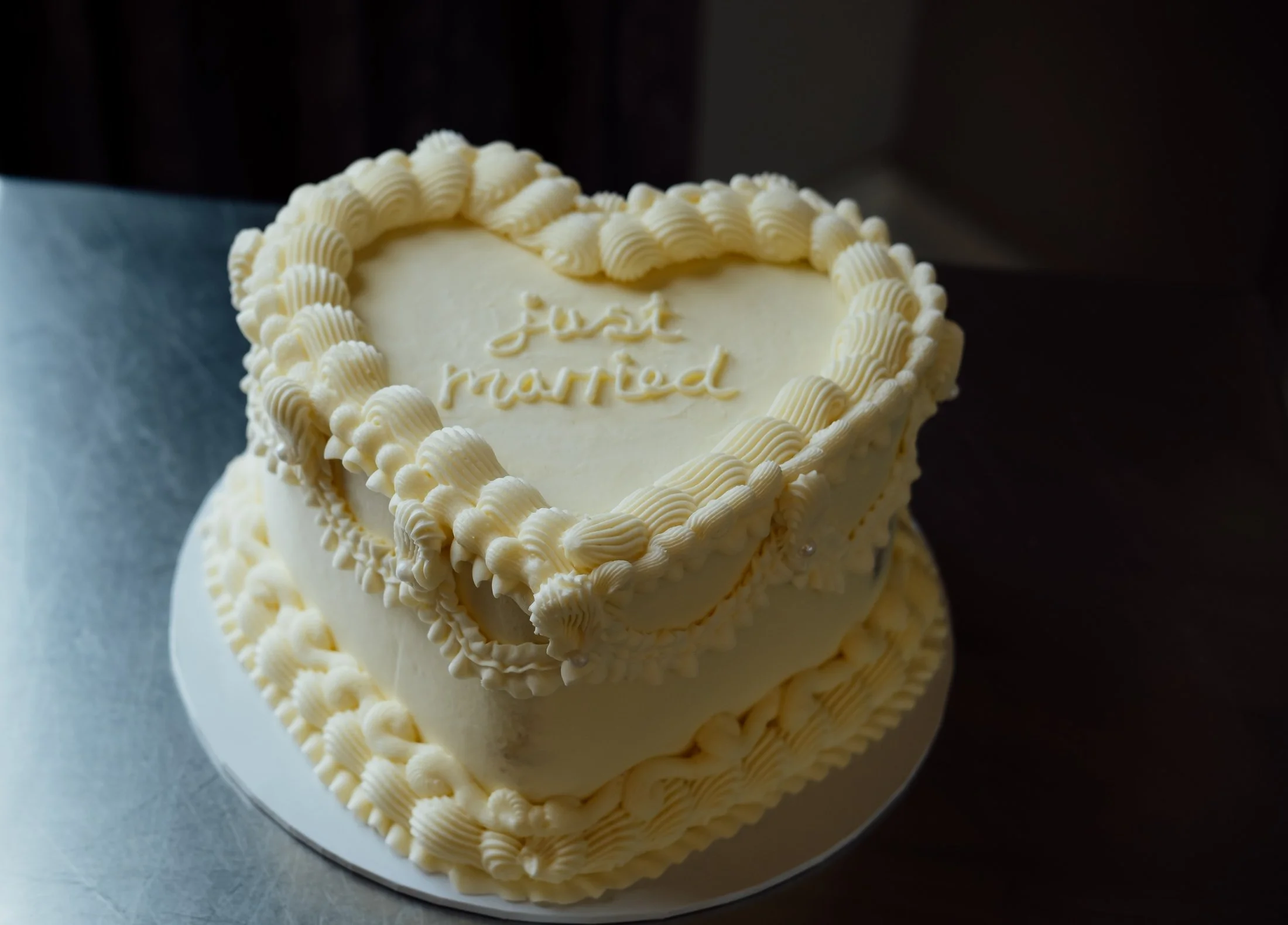 Cream-colored, heart-shaped wedding cake with white icing and decorative piping. The top of the cake has a message that reads "Just Married" in cursive writing.