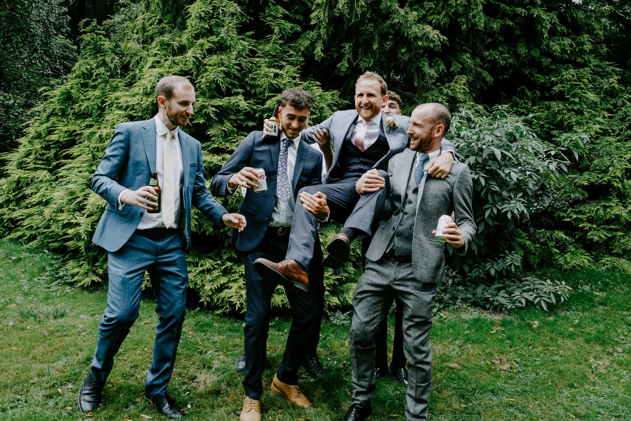 Group of six men in suits celebrating outdoors, one being carried on others' shoulders, holding drinks, in front of green bushes.