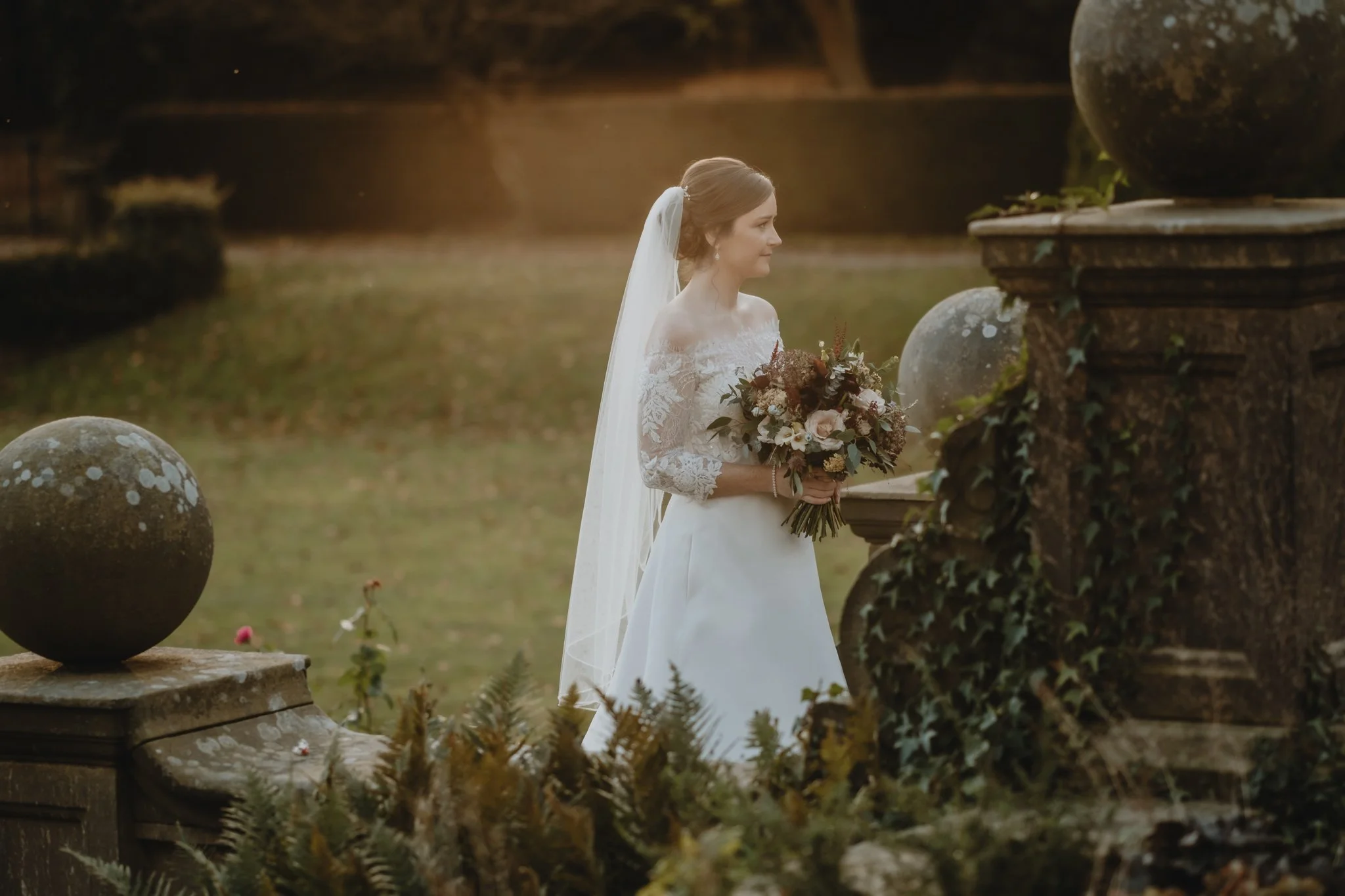A bride in a white lace wedding dress holding a bouquet of flowers, standing outdoors near stone balustrades and greenery, with a softly lit background.