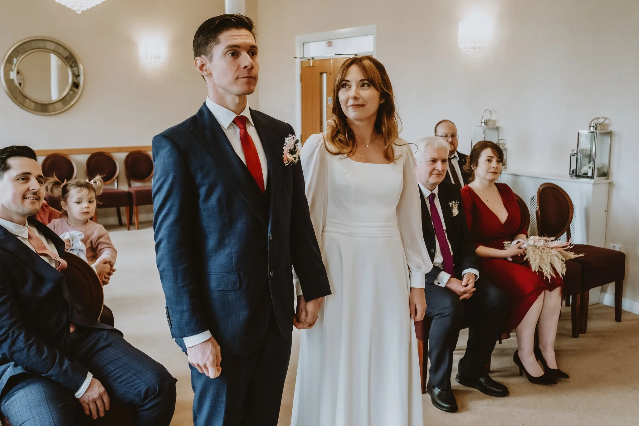 A wedding ceremony with the bride and groom standing, surrounded by seated guests in an indoor setting.