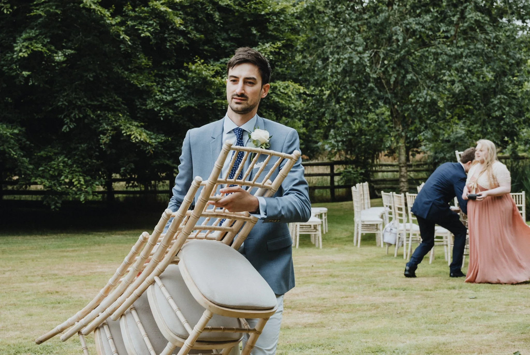 A man in a light blue suit holding a chair outdoors during a wedding ceremony, with a woman in a peach-colored dress and a man in a dark suit kissing in the background.