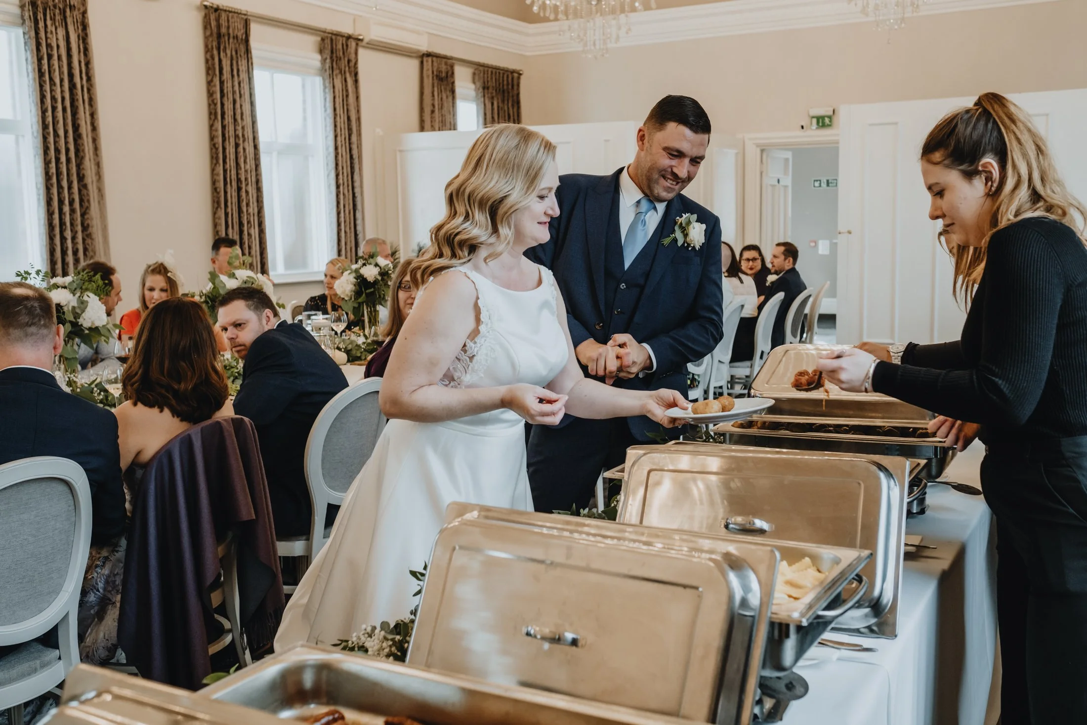 A bride and groom smiling while serving themselves food at a wedding reception, with guests seated at decorated tables in the background.