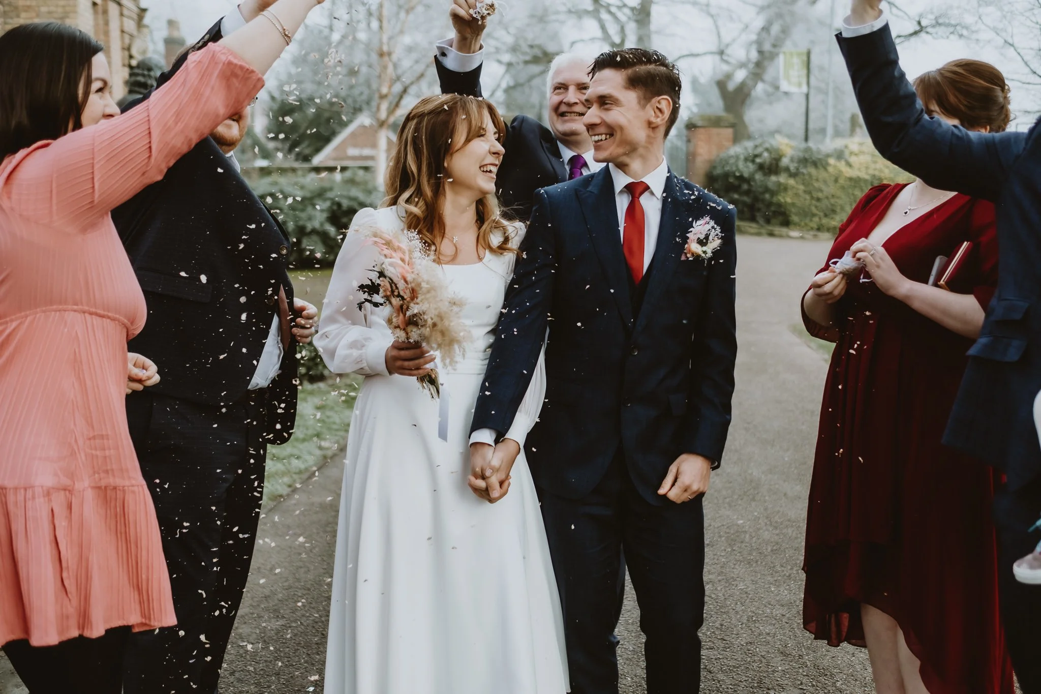 Newlywed couple holding hands and smiling as friends and family celebrate with cheers and confetti outdoors.