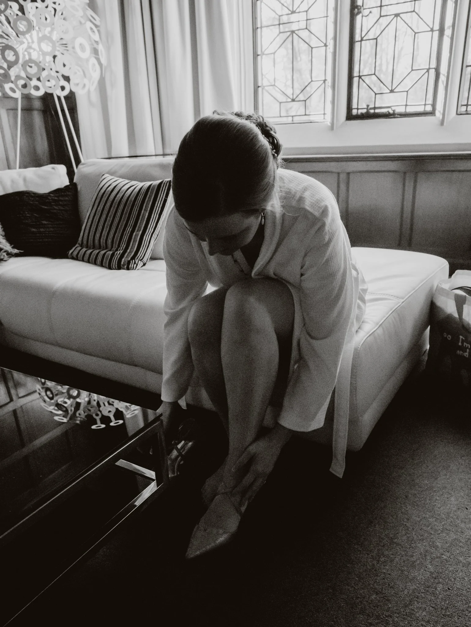 A woman in a white robe is sitting on a couch, bending forward to put on her shoes. The room has a window with stained glass and sheer curtains, with decorative pillows on the couch and a lamp with a circular patterned lampshade.