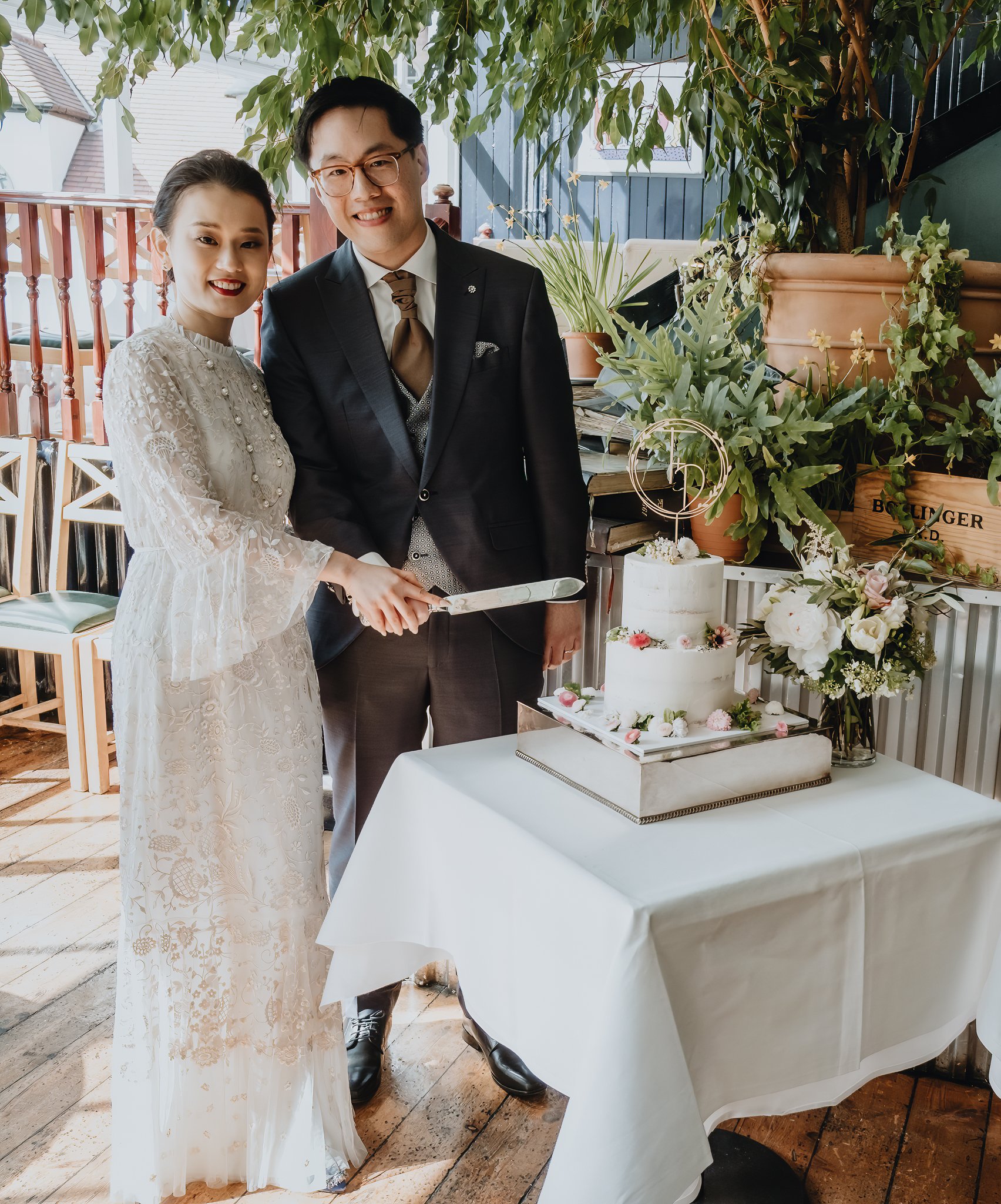 A bride and groom cutting a wedding cake on a table decorated with flowers, under a green leafy canopy.