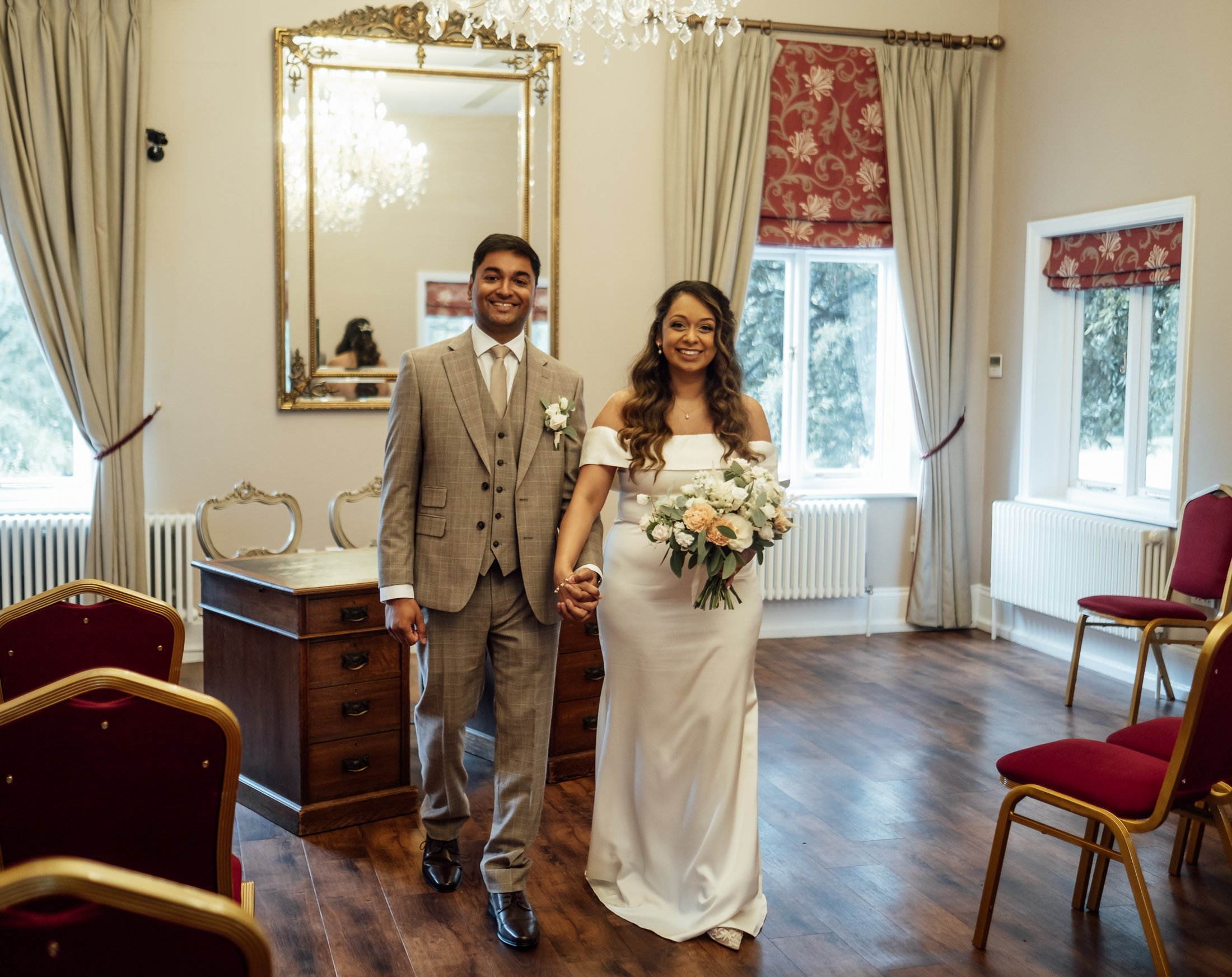 A couple in wedding attire standing in a decorated room, holding hands and smiling, with white curtains, red and gold chairs, and a large mirror in the background.
