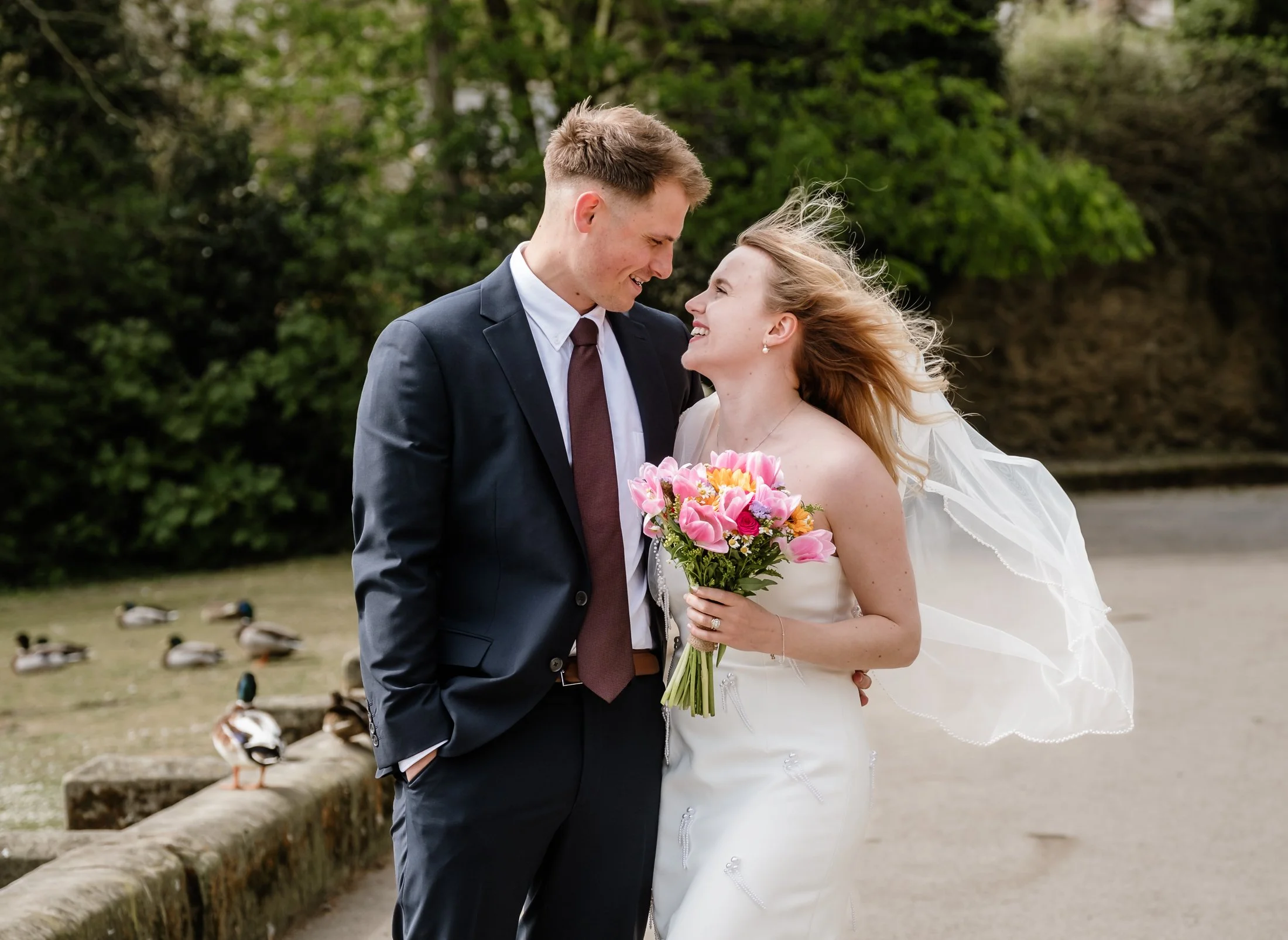 A bride and groom smiling at each other outdoors near a pond with ducks, with trees in the background.