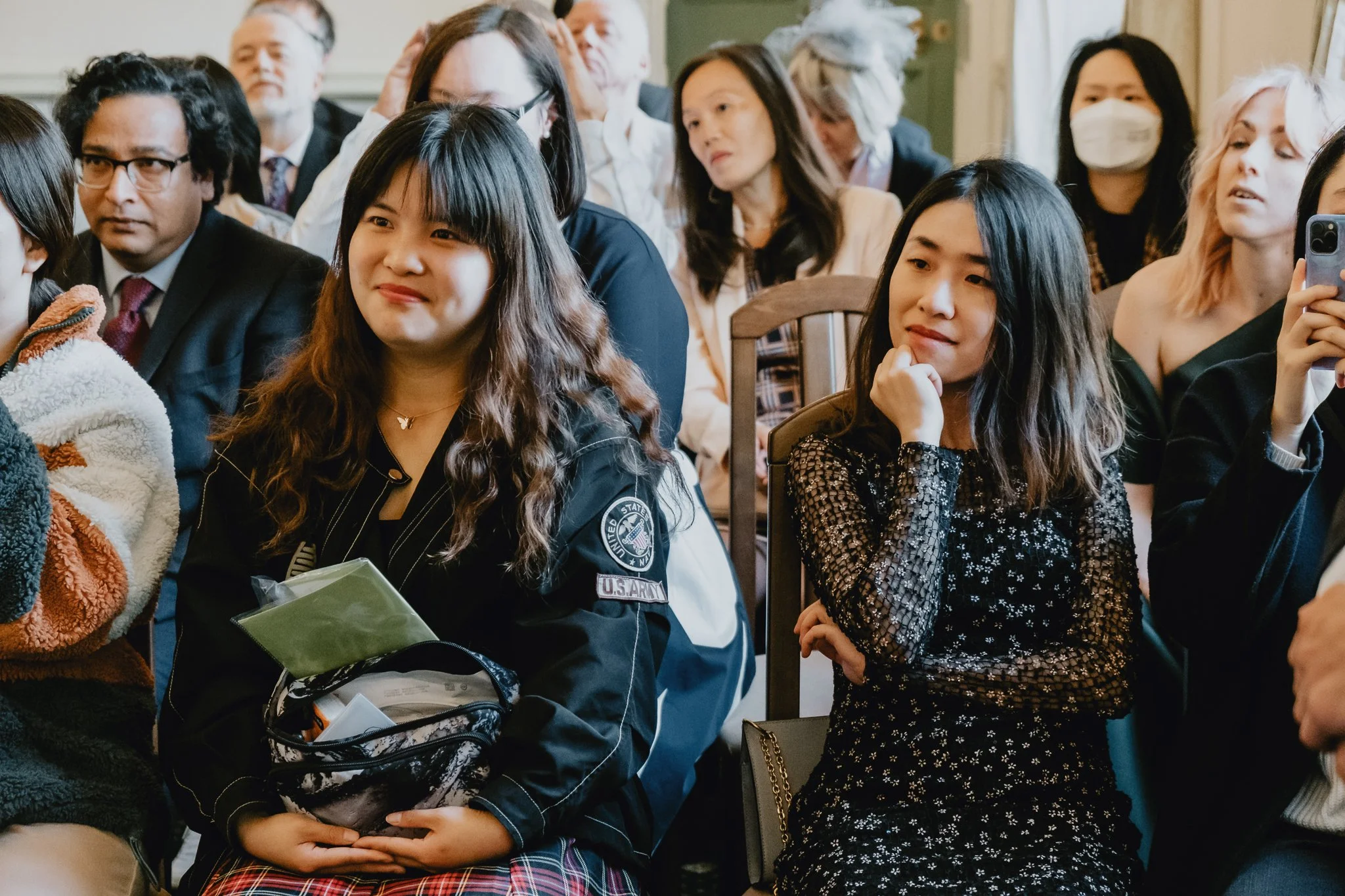 A diverse group of people seated and watching an event, including two young women in the foreground, one with long wavy hair wearing a U.S. Army jacket, and the other with shoulder-length hair in a black floral dress.