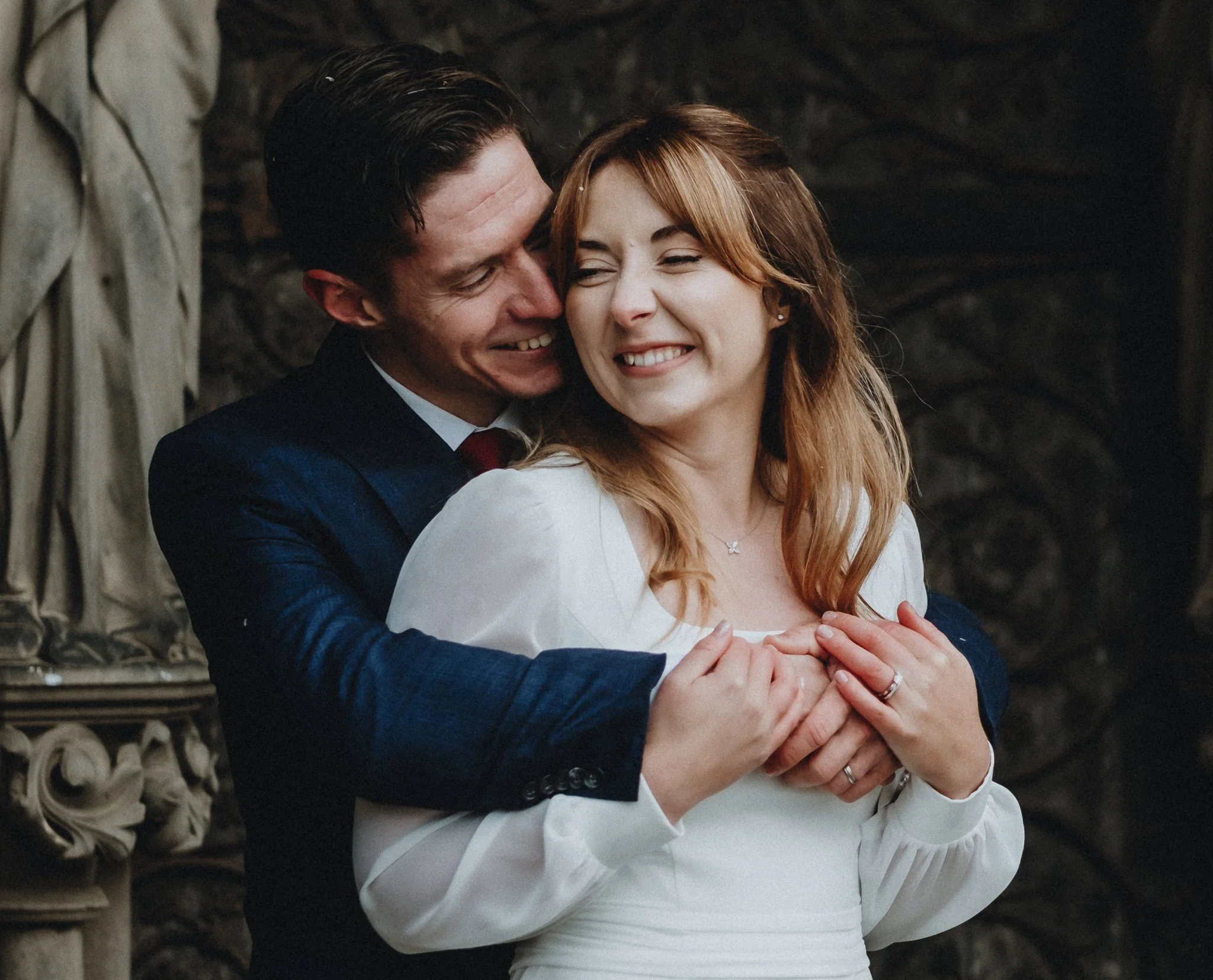 A couple sharing a joyful hug, smiling, with the man hugging the woman from behind and both holding her hands, in an indoor setting with ornate architectural details.