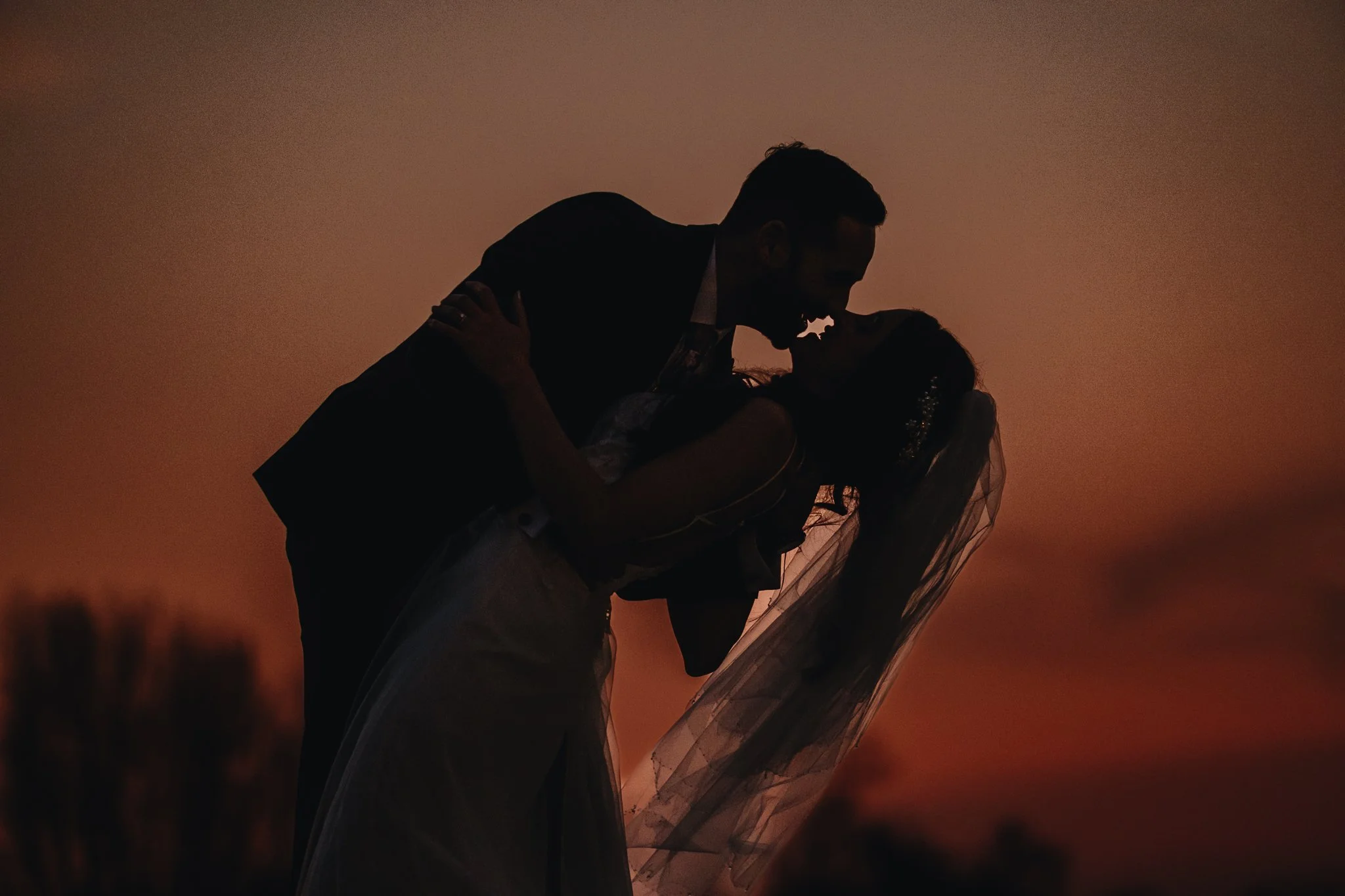 Silhouetted couple sharing a kiss during sunset, with the man leaning over the woman, both dressed in formal attire.