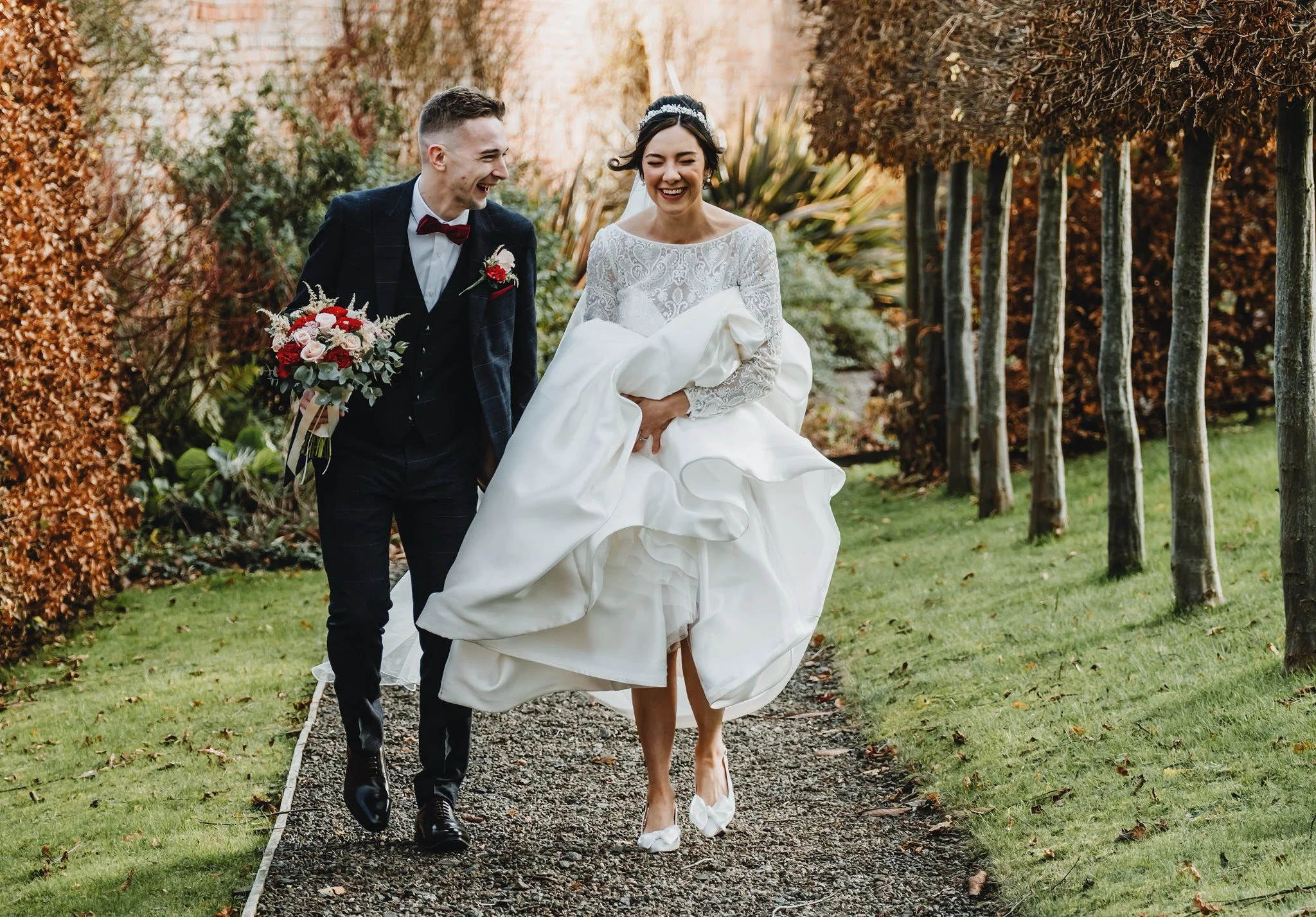 A bride in a white wedding gown and a groom in a dark suit walk outdoors on a garden path, smiling and laughing together, with trees and autumn foliage in the background.